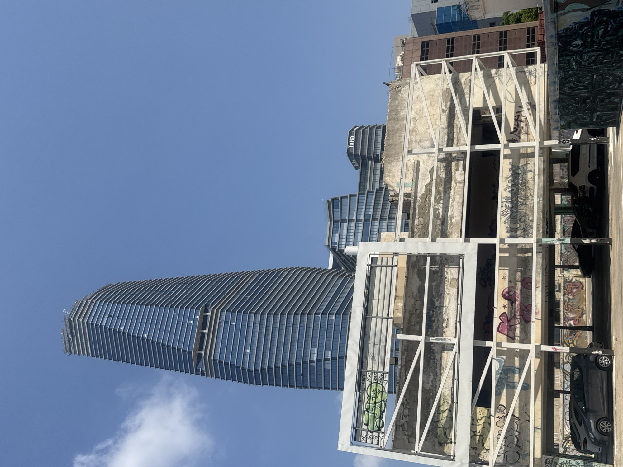 The image shows a street view in Tel-Aviv, Israel, under a clear blue sky with scattered white clouds. In the foreground, an unfinished or partially demolished multi-story structure, possibly a parking garage, is visible. Its concrete walls are covered in graffiti, and exposed metal beams form its framework. Several cars are parked on the ground level of this structure. Behind it, a sleek, modern skyscraper with a glass facade dominates the skyline. To the right of the skyscraper, other contemporary buildings, including one with the signage "ToHa", are present, indicating an urban commercial or residential area. The contrast between the dilapidated foreground structure and the modern architecture behind highlights urban development and potentially ongoing construction or renewal in the city.