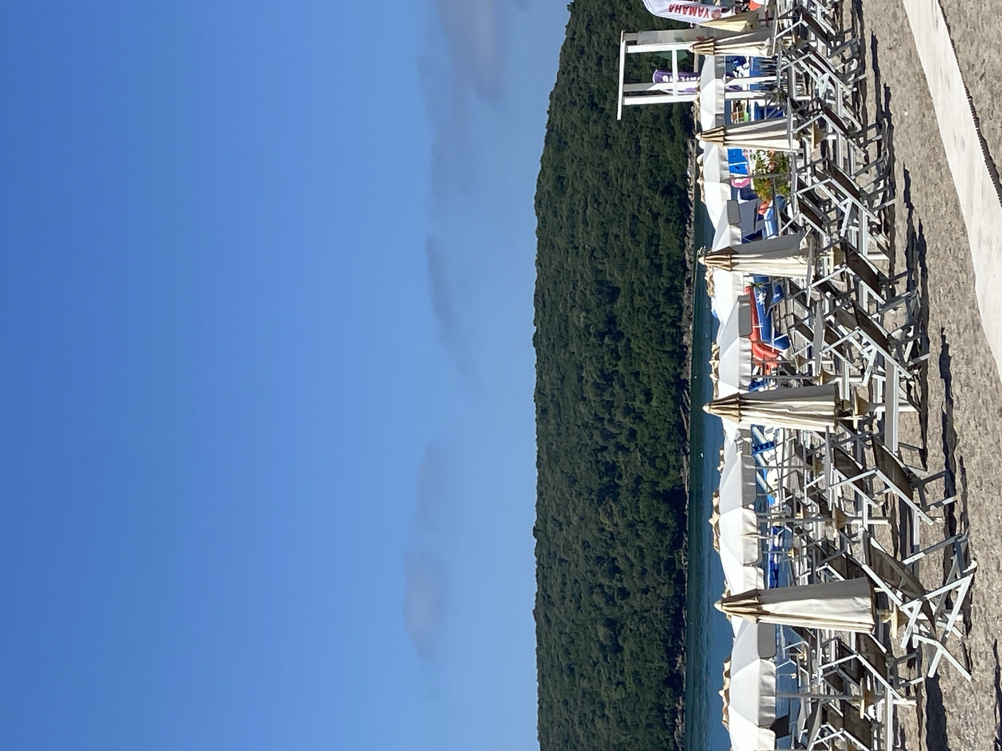 This is a photograph taken from a beach in Prijevor, Montenegro. In the foreground, rows of empty lounge chairs and closed beach umbrellas are set up on the sand. Beyond the chairs, the blue water of the sea is visible, with a densely forested green hill forming the backdrop. The sky is a clear, bright blue with a few wispy clouds. There is a white lifeguard stand on the right side of the image, and a person wearing a white shirt with "YAMAHA" printed on it can be seen standing near it. The overall scene depicts a serene beach day with no people actively engaged in beach activities.