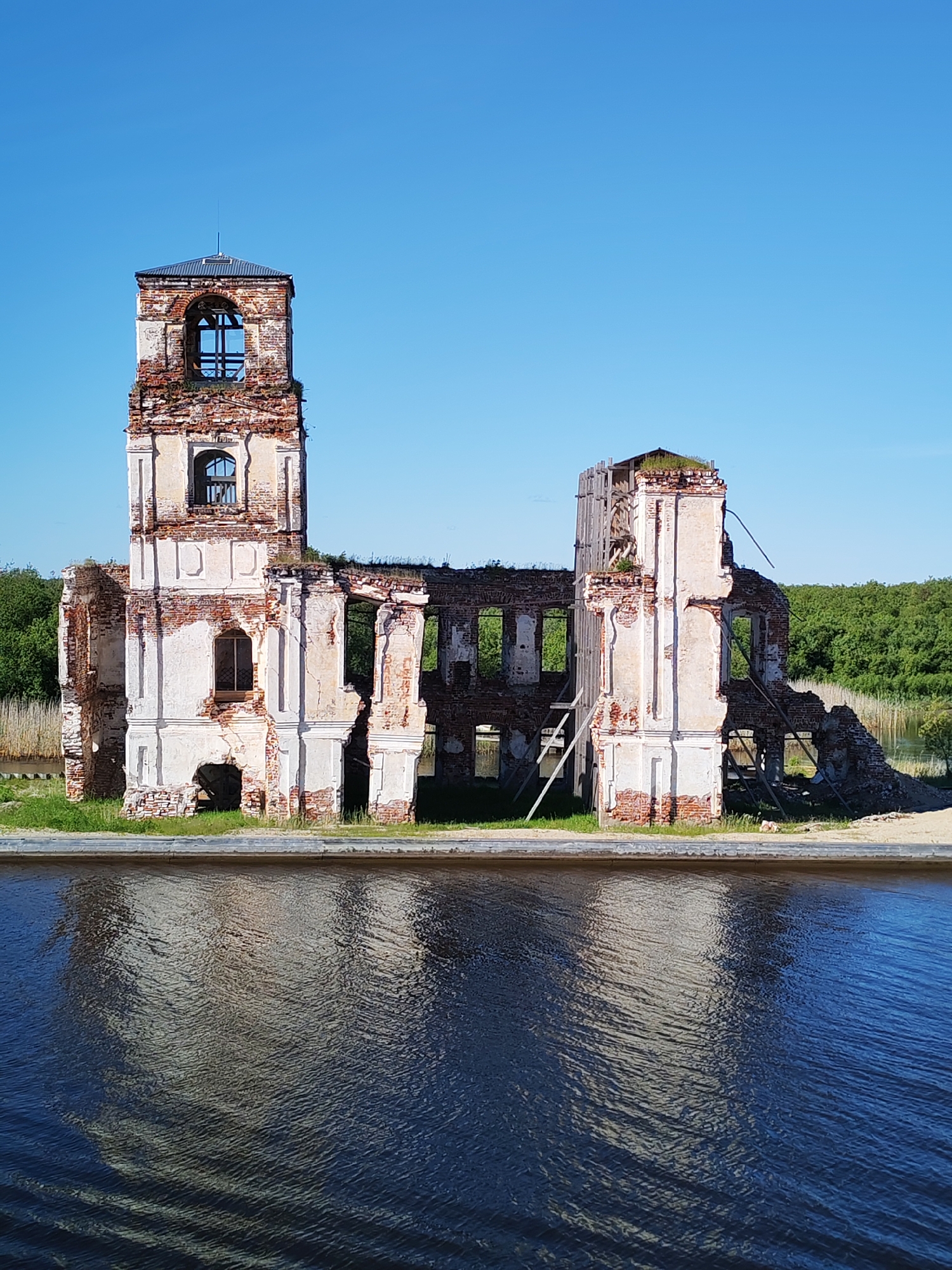 This vertical shot captures the evocative ruins of a historic building in Krokhino, Russia, standing by a body of water under a pristine blue sky. The structure, likely a former church or manor, is severely dilapidated, showcasing crumbling brickwork and remnants of white plaster.
The most prominent feature is a tall, partially intact tower on the left side of the ruin, capped with a dark metal roof and featuring arched window openings. The rest of the building is largely a skeletal shell, with extensive portions of its walls and roof missing, revealing open spaces and the internal structure. Exposed red brick is a dominant material, contrasting with the faded white plaster that still clings to some architectural elements. Scattered wooden beams and what appear to be temporary scaffolding or supports are visible within the collapsed sections, particularly on the right side of the ruin. Green grass and sparse vegetation grow around the base of the ruin.
In the foreground, a calm body of water, possibly a canal or river, reflects the ruined edifice with dark, rippled patterns. A narrow strip of land separates the building from the water's edge. The background features a dense line of lush green trees and reeds, indicating a natural landscape surrounding the site. The sky is a vivid, cloudless blue, suggesting bright and clear weather, likely mid-day or afternoon, given the strong illumination. There are no people or discernible text visible in the image. The scene conveys a sense of historical decay and serene abandonment.