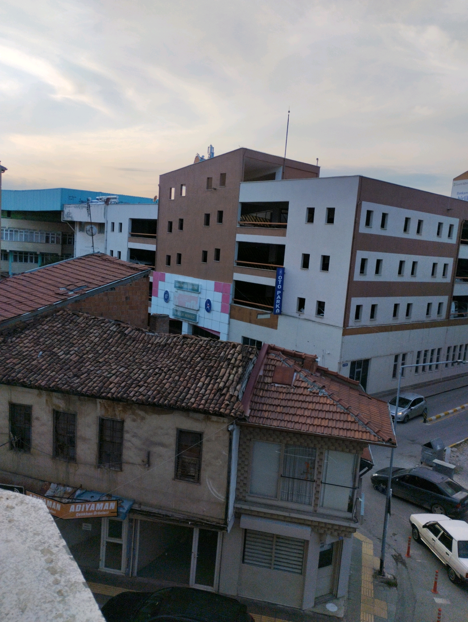 This aerial view captures a street scene in Çorum, Turkey, focusing on a cluster of buildings. In the foreground, an older, two-story building with a red tile roof dominates the left side. Its facade is weathered and shows signs of age, with several windows visible. A sign for "ADIYAMAN Çelikhan Ürünleri" (Adıyaman Steel Products) is mounted above the ground-floor entrance.
Adjacent to this older building and slightly behind it, a multi-story parking garage stands out. This modern structure has a white and brown facade with numerous rectangular windows. A vertical blue sign with white text reads "OTO PARKA." Further back, another, taller building with a similar architectural style is visible, featuring multiple floors and a repeating pattern of windows. A large billboard-like structure with pink and white panels and text is attached to its side.
On the street level, several cars are parked and driving. A silver hatchback is seen turning on the road. A dark-colored sedan is parked on the side, and a white sedan is also visible. Traffic cones are placed along the roadside. The sky above is overcast with some streaks of clouds, suggesting it might be late afternoon or early morning. The urban environment appears to be a mix of older and newer constructions, indicative of a developing city.