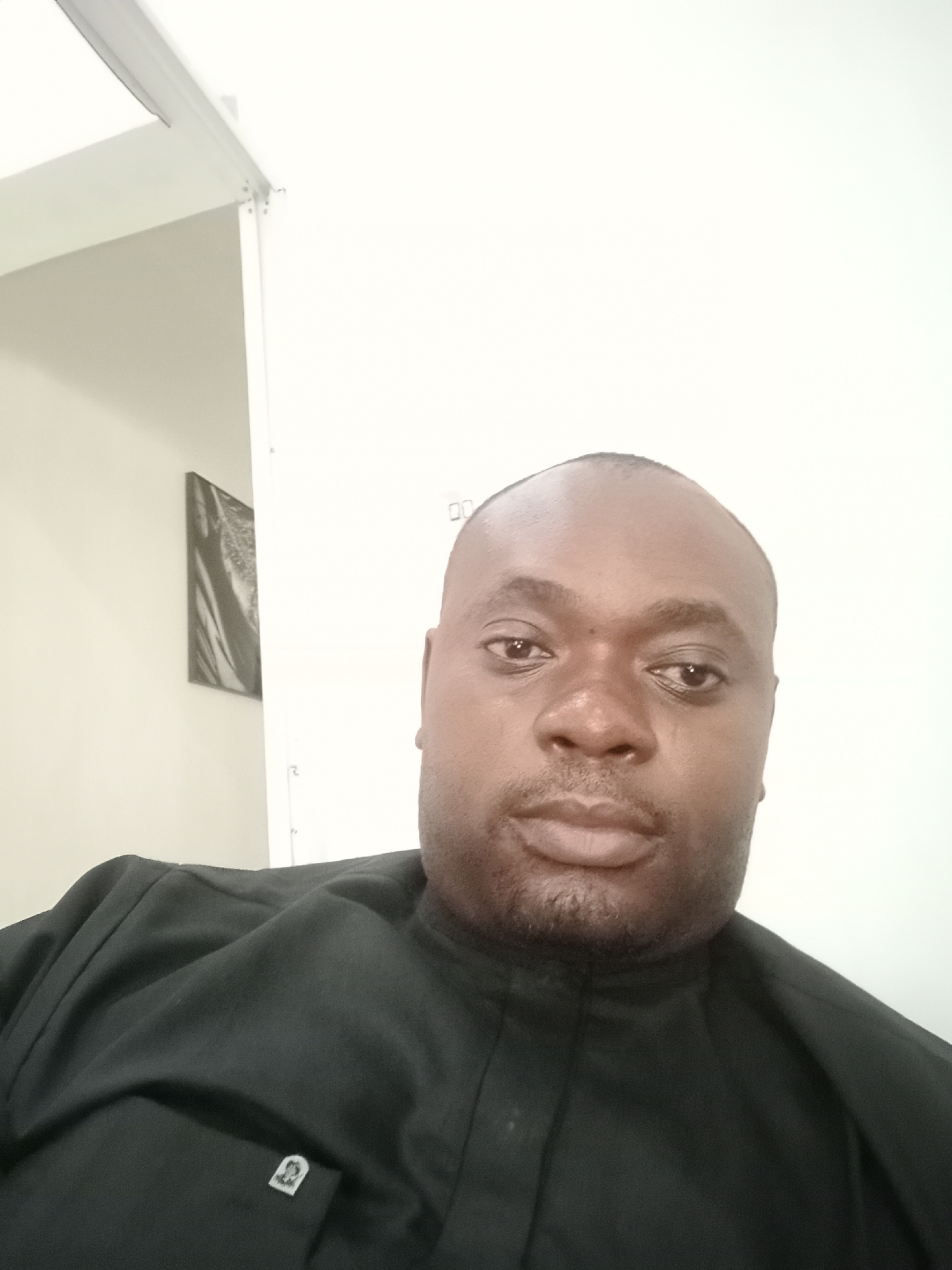 A man of African descent, likely in his 30s or 40s, is featured in a close-up, head-and-shoulders portrait. He has dark skin, a bald or closely shaved head, and a short beard or stubble. He looks directly into the camera with a neutral expression.
The man is wearing a black, traditional-style garment, possibly an Agbada, with a high, simple collar. A small, square lapel pin with an indistinct silver-colored emblem or crest is visible on the left chest area of his attire. No discernible text is visible on the pin or elsewhere in the image.
The setting appears to be indoors in Gwarinpa, Nigeria. The background consists primarily of plain white walls, suggesting a brightly lit interior space. To the left and behind the man, a dark, framed piece of artwork or photograph with abstract patterns hangs on a light wall. In the upper left quadrant, a white ceiling and part of a light fixture or structural beam are visible. A white vertical partition or doorframe is also partially visible on the right side of the frame. The overall lighting is bright and even, consistent with an indoor environment during the daytime.