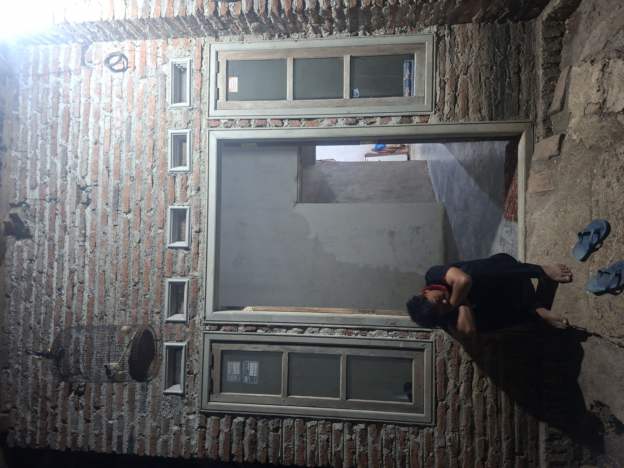 The image depicts an outdoor scene at night in Tongaswetan, Indonesia. A person is sitting on the ground, hunched over with their head resting on their arms. They are wearing a black t-shirt with a red collar and black pants. Two pairs of blue flip-flops are on the ground near the person.
The backdrop is a brick wall with several windows and ventilation bricks. A birdcage with two birds is hanging from the wall. The scene appears to be in front of a doorway, with a glimpse of an interior space visible through it. The lighting suggests an artificial light source is illuminating the area from the right. The ground is uneven and appears to be made of dirt and rough stones.