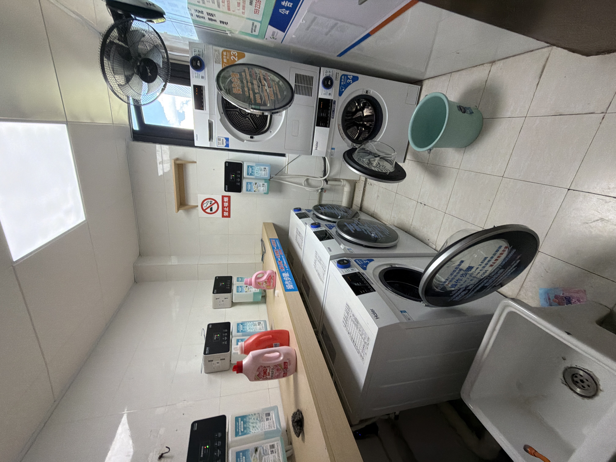 The image depicts the interior of a communal laundry room in Jiangjin, China. The room is tiled with white and light-colored tiles on the walls and floor. Along the back wall, a wooden shelf holds several bottles of laundry detergent, two of which are pink and red. Below the shelf, mounted on the wall, are several control panels for the laundry machines, some with small screens displaying information. A sign with a cigarette symbol and Chinese characters indicating "No Smoking" is also visible on the wall.
Dominating the center of the image are several washing machines and dryers. A stacked unit features a dryer on top and a washing machine below. Both the dryer door and the washing machine door are open, revealing the drums inside. The dryer drum displays a circular sticker with Chinese text. Next to this stacked unit, another washing machine is visible with its door open. To the left, two more washing machines are partially visible.
In the foreground, a large white sink with a drain is present. A light blue bucket sits on the floor near the washing machines. A ceiling fan is mounted high on the wall to the right, and a large window with a view of blue sky and clouds is visible in the background. The lighting in the room appears to be from overhead fluorescent panels. The overall impression is that of a functional, albeit slightly cluttered, public laundry facility.