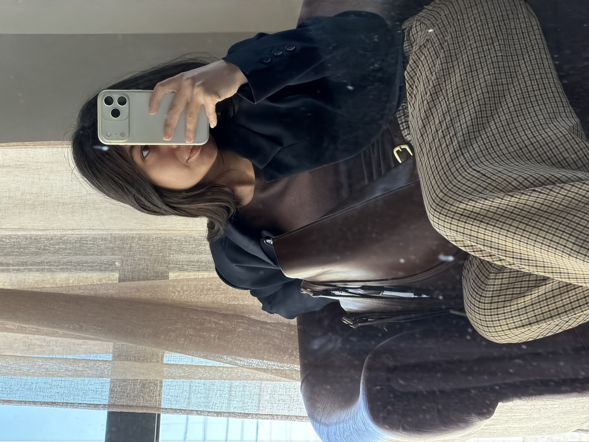 A person is taking a selfie in a mirror. The person is wearing a black blazer over a brown top, with plaid pants and a brown purse with a gold buckle. The person is holding a white smartphone to take the photo. The background shows a window with tan blinds. The reflection implies an interior setting with a chair. The city or precise location is not discernible from the provided image.