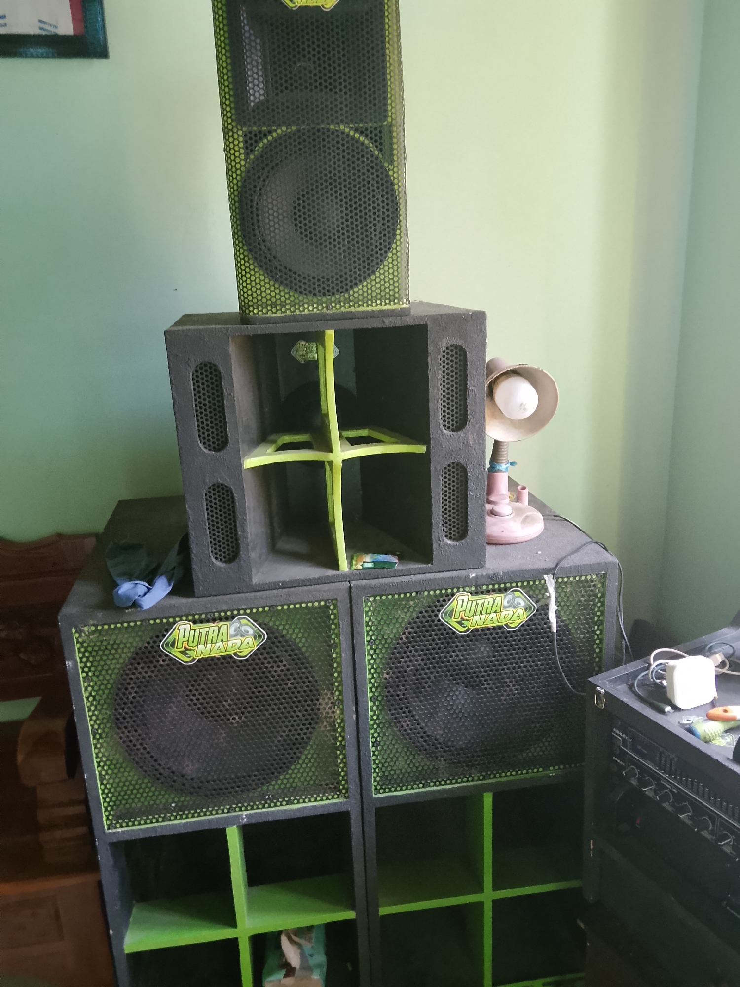 The image shows a stack of large black speakers with green accents in a room with pale green walls. The speakers are of various sizes, with the largest at the bottom and progressively smaller ones stacked on top. The bottom two speakers are identical, each featuring a large circular speaker grille with a green border and a green sticker logo that reads "PUTRA NADA". Above these are two more speakers, one a rectangular cabinet with visible internal green shelving, and the top speaker is a tall, narrow unit with a visible speaker grille. To the right of the speaker stack, a small pink desk lamp with a white bulb is present on top of a speaker. A pile of electronics, including what appears to be a rackmount amplifier or mixer, is visible to the far right. A blue cloth is draped over the speaker to the left. The environment appears to be indoors, likely a room or storage area. The overall impression is of a sound system setup. The image was taken in Tawangsari, Indonesia.