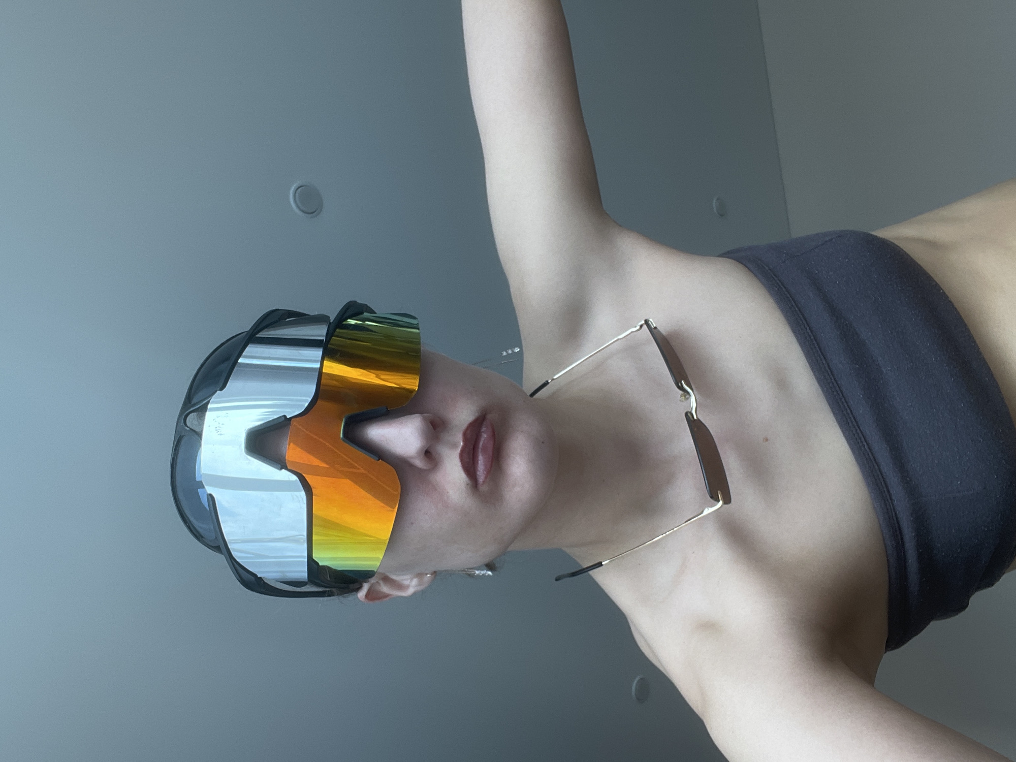 A woman wearing a black sports bra and futuristic sunglasses is in an indoor environment. She is also wearing glasses around her neck. The background consists of a plain, light-colored wall with circular fixtures. There is no verifiable evidence to confirm the location is in Подшивалово, Russia.