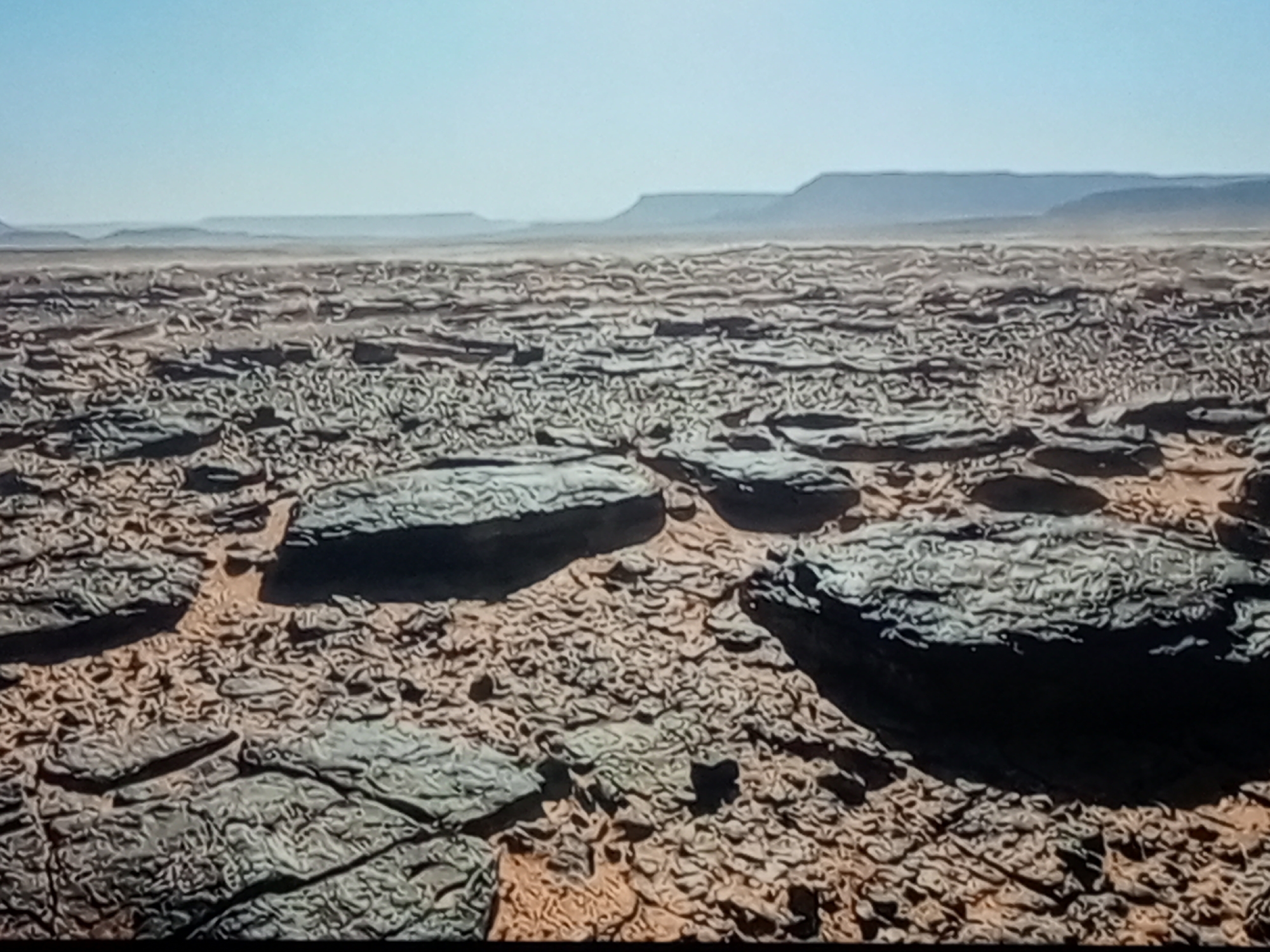 This is a wide shot of a rocky desert landscape under a clear sky. In the foreground, large, dark, irregular rocks are scattered across a surface of smaller, lighter-colored, eroded rocks and sand. The texture of the ground is rough and uneven, with many small, fragmented stones. In the middle ground, the terrain continues with a similar rocky texture, stretching towards a hazy horizon. Distant, flat-topped mountains or mesas are visible in the far background, silhouetted against the bright, pale blue sky. The scene is arid and desolate, with no visible vegetation or signs of life. The lighting suggests it is daytime, with shadows indicating the direction of the sun.