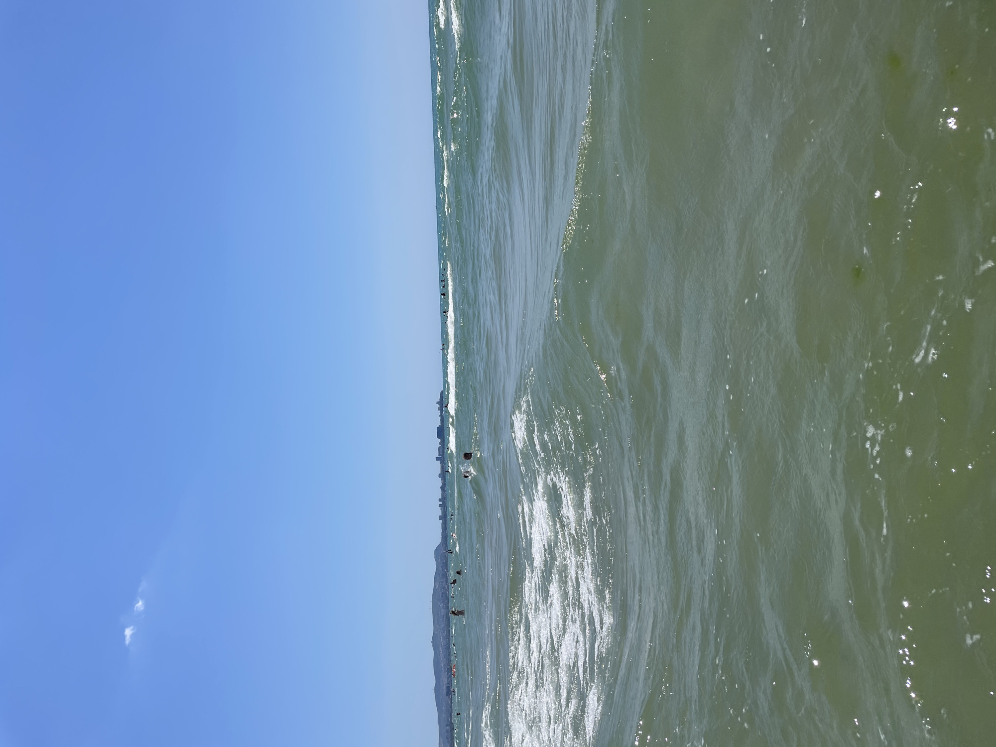 A wide-angle, eye-level shot captures a sun-drenched beach scene in Anapa, Russia. The ocean fills the foreground, with clear, green-tinted water showing gentle waves and white foam. The sun glints off the water's surface, creating a sparkling effect. In the middle ground, several people are visible swimming and wading in the sea. The background features a distant coastline with a town and hills under a clear blue sky with a few wispy white clouds. The overall impression is one of a pleasant summer day at the seaside.