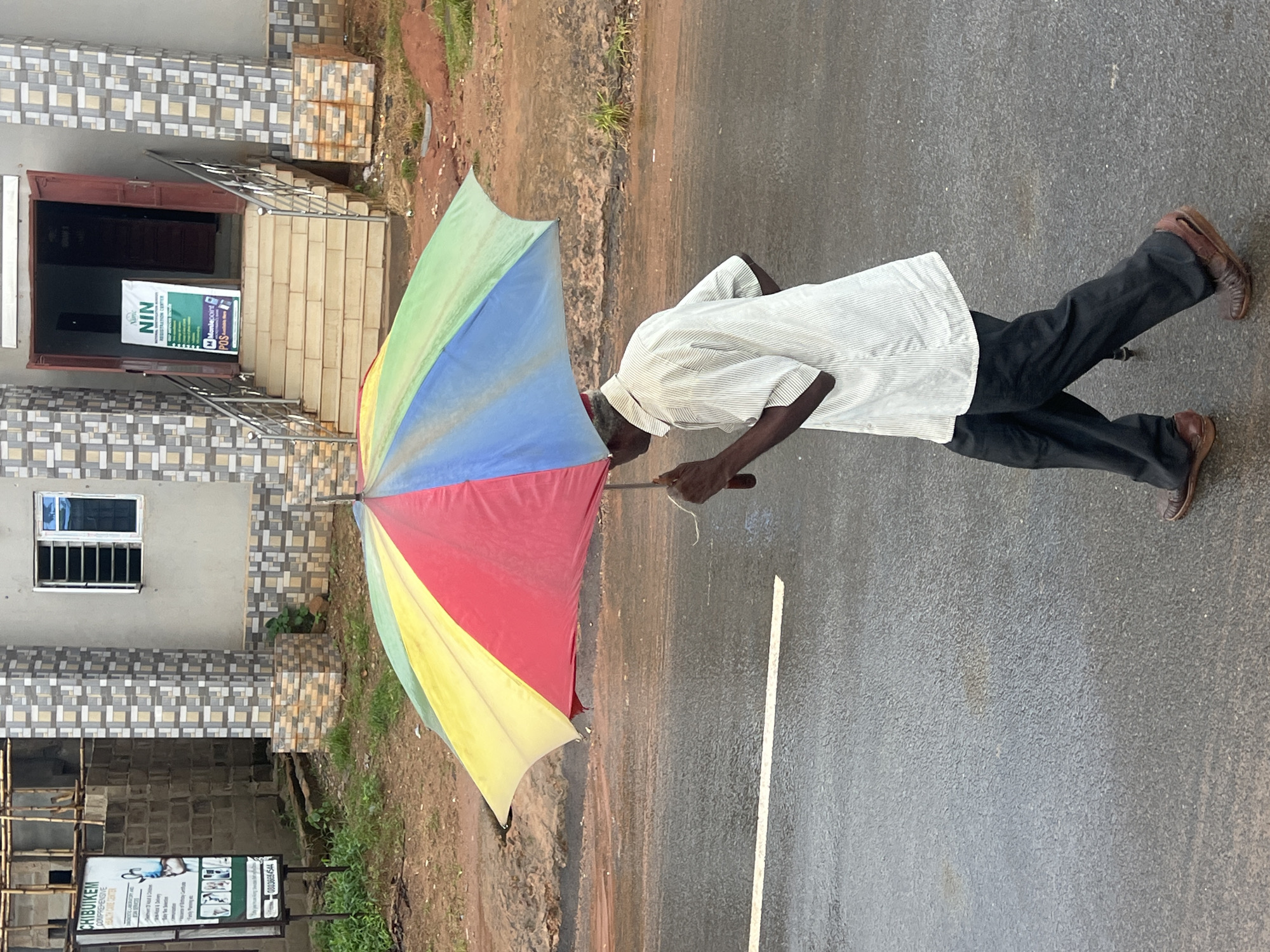 The image shows a person holding a colorful umbrella in Nkpor, Nigeria. The person is wearing a striped white shirt and dark pants, with brown shoes. They are standing on a wet asphalt road, suggesting it might be raining or has recently rained. The umbrella is open and displays a rainbow of colors: green, blue, red, and yellow. The scene is set in front of a building with a tiled facade and a set of stairs leading up to an entrance. A sign with the text "NIN" is visible near the entrance, possibly indicating a National Identification Network office. Another sign with text that includes "CHIBUIKEM" and a phone number "08036654544" is visible on the left side of the image, near a construction area. The overall impression is one of a typical day in a Nigerian town, possibly during the rainy season. The lighting suggests it is daytime.