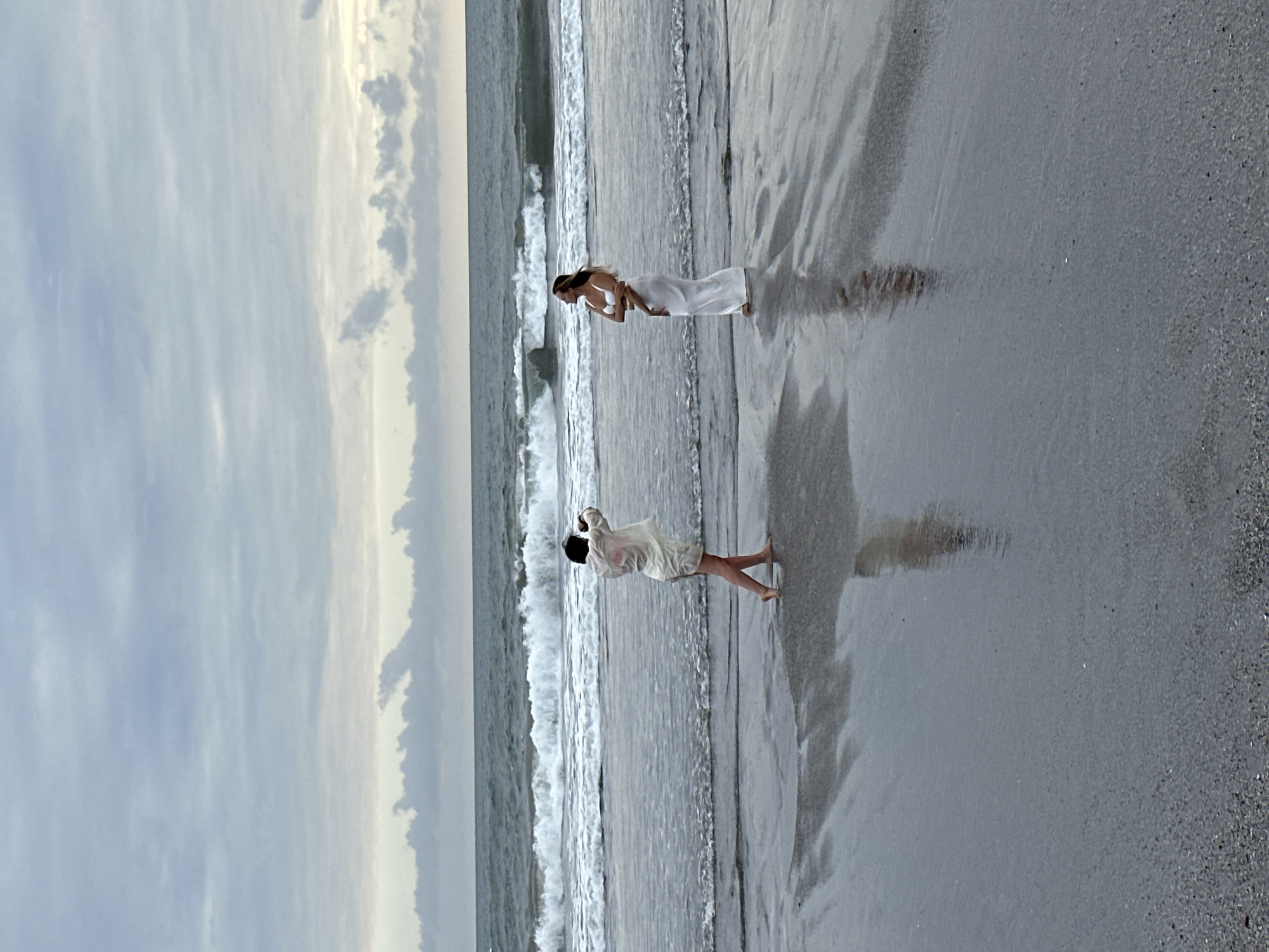 The image depicts two women on a beach in North Kuta, Indonesia, during overcast weather. In the foreground, a woman is standing in shallow water on the wet sand, facing towards the ocean and holding a camera up to take a photo. She is wearing a light-colored, loose-fitting outfit. Further back, another woman is standing in the water, facing away from the camera and looking towards the sky. She is wearing a long, white dress. The ocean has moderate waves, and the sky is filled with heavy, gray clouds. The wet sand reflects the figures of the two women and the waves. The overall scene conveys a sense of calm and serene natural beauty, with the two women engaged in photography and contemplation by the sea.