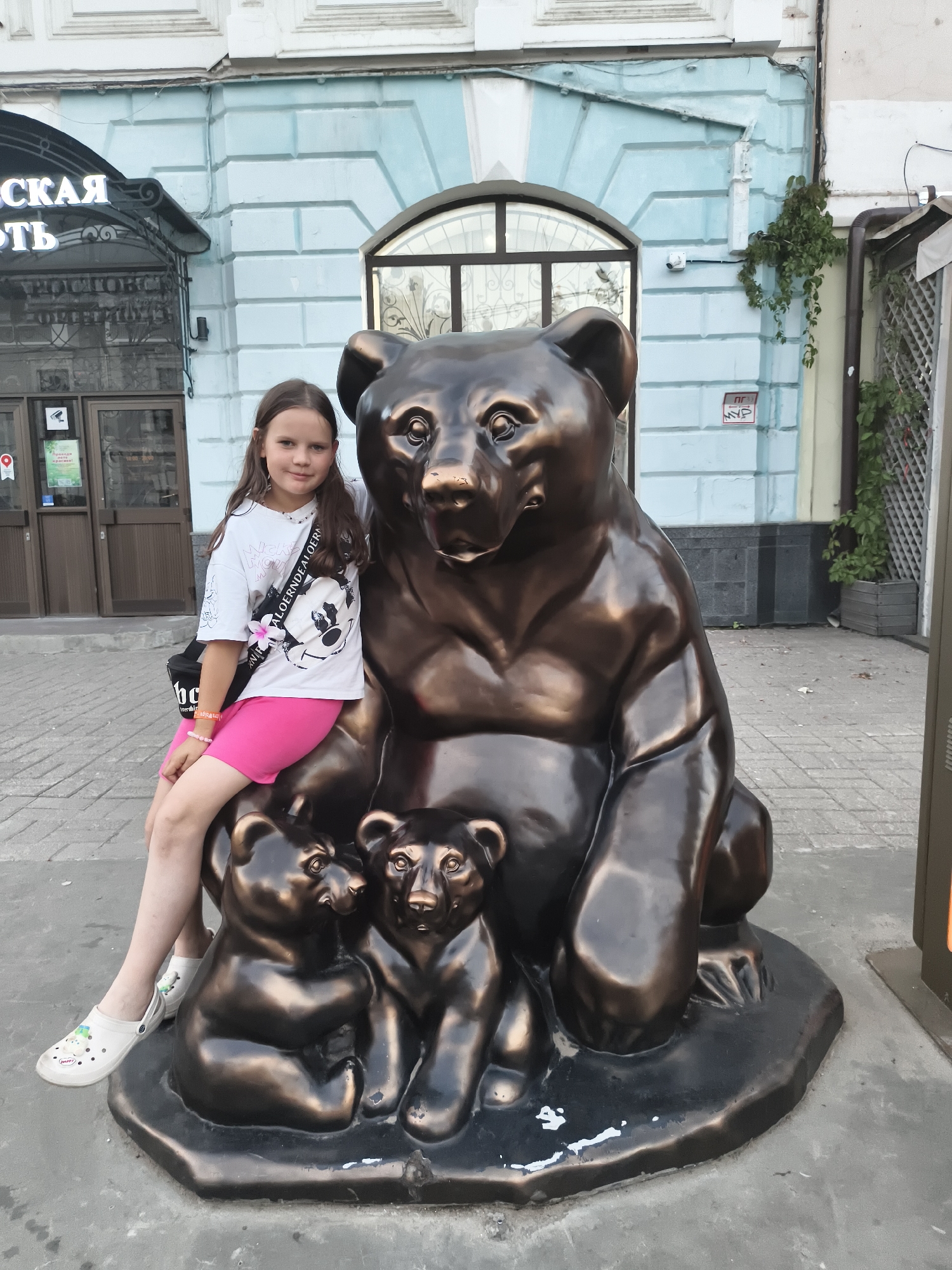 In Yaroslavl, Russia, a young girl with brown hair is photographed sitting on the lap of a large bronze bear statue. She is wearing a white t-shirt with a graphic print and pink shorts. The bear statue is part of a sculpture that also includes two smaller bear cubs sitting at its feet. The girl is smiling at the camera. The background shows a building with light blue walls and architectural details, along with part of a shop entrance. The setting appears to be an outdoor public space, likely a street or pedestrian area.