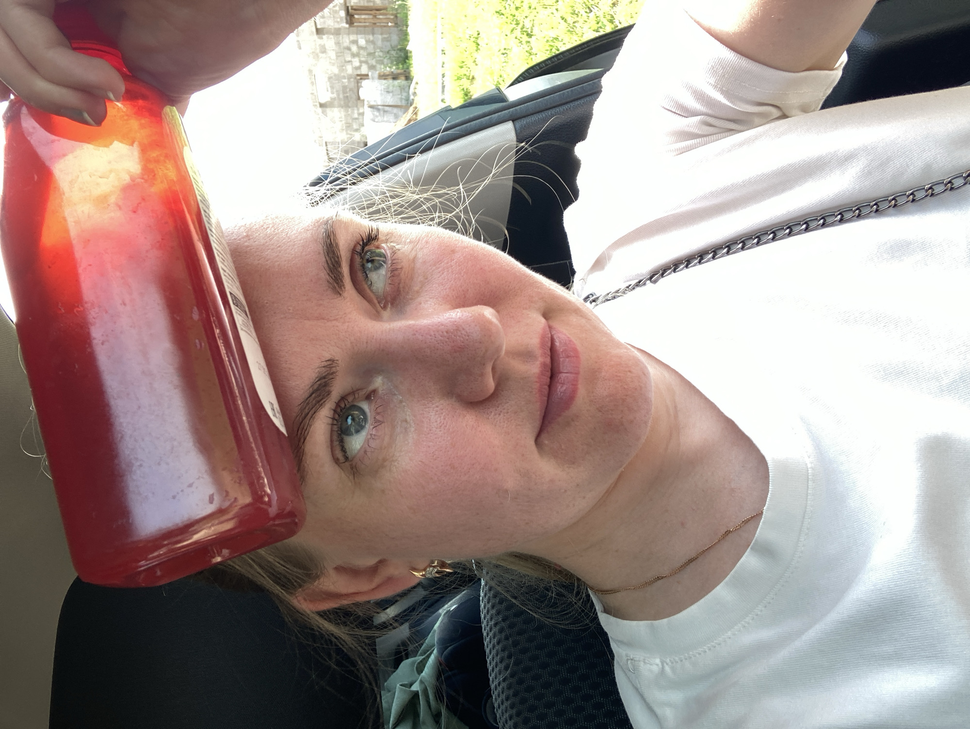 A woman is pictured in a car, holding a red bottle over her forehead. She appears to be looking off to the right of the frame. Her hair is blonde and she is wearing a white t-shirt. The background outside the car shows a sunny day with green grass and what appears to be a construction site with brick buildings. The exact location in Fokino, Russia is not discernible from the image.