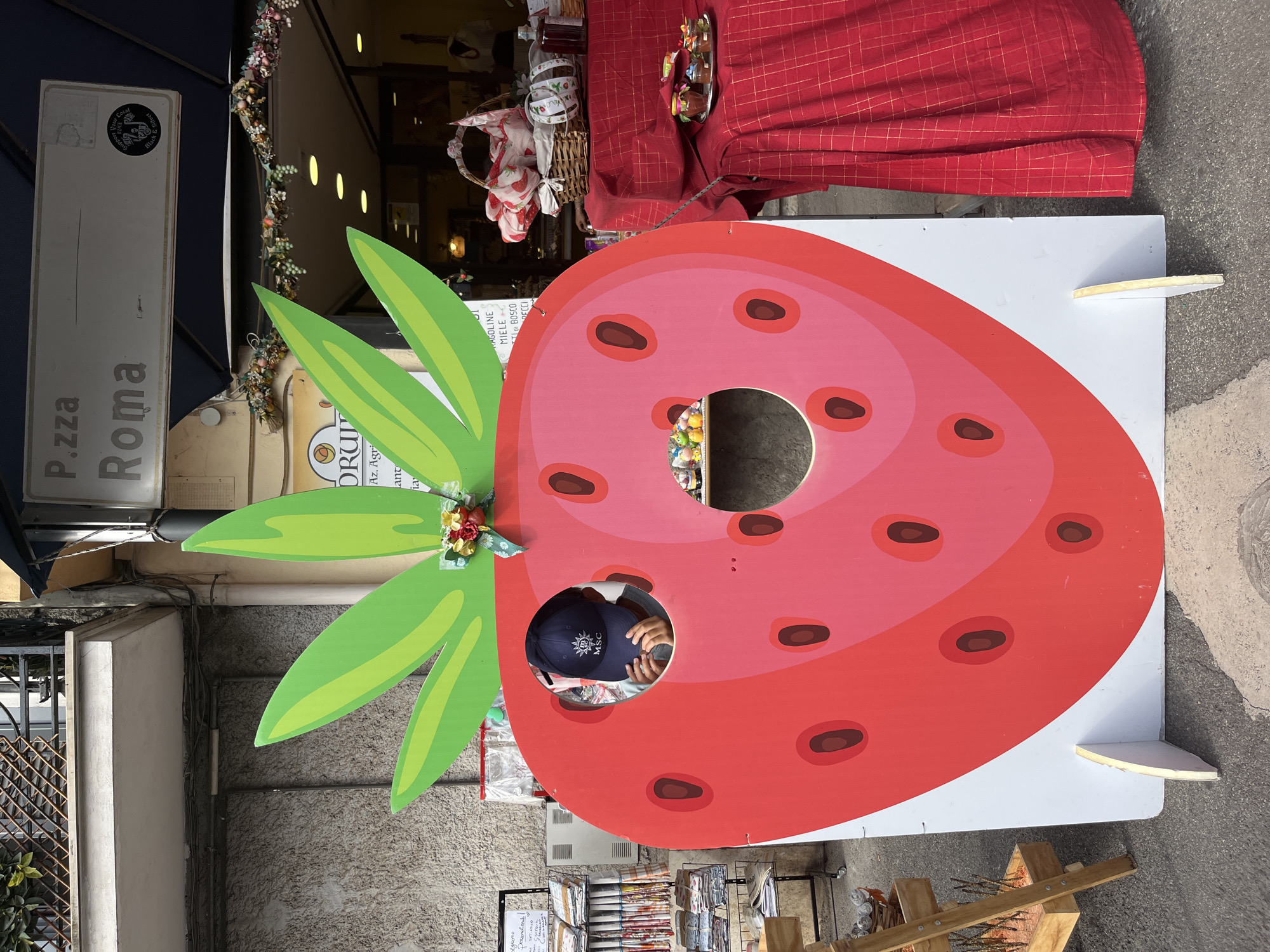 The image depicts a street scene in Nemi, Italy, featuring a large, flat, red strawberry cutout with two holes, one for a face and another lower down. A person wearing a dark blue baseball cap with a white logo is peeking through the upper hole, their hands visible. The strawberry cutout stands on a white base. To the right, a table draped with a red tablecloth displays various jars and baskets of goods, possibly local produce or souvenirs. In the background, a building with a sign reading "P.zza Roma" is visible, along with a shopfront and hanging decorations. To the left, shelves filled with folded textiles and other items are present. The ground is paved.