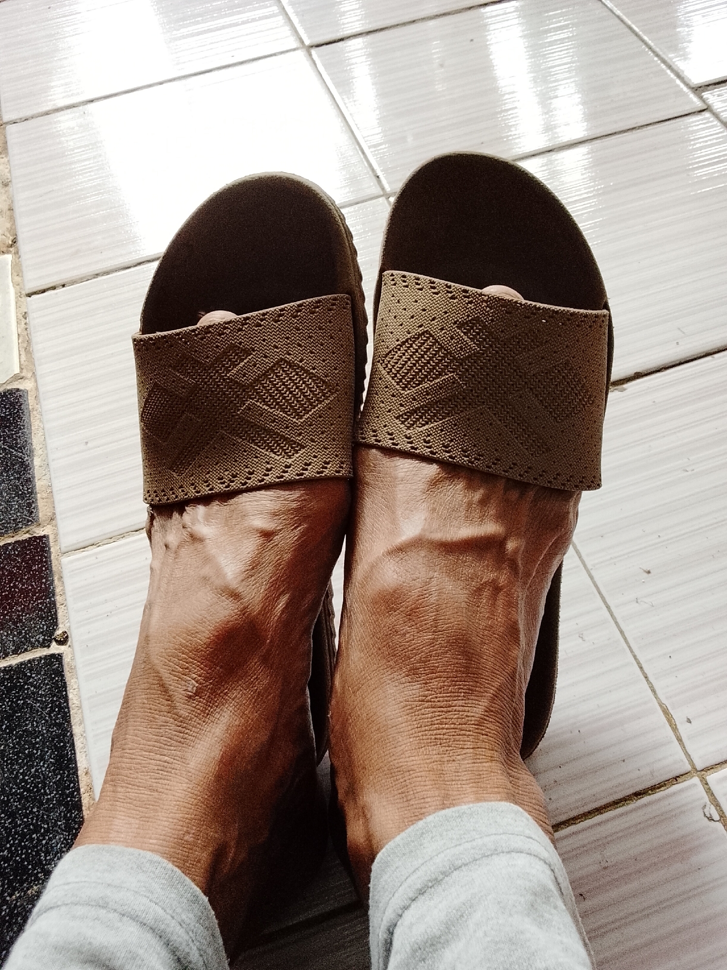 The image shows a close-up, top-down view of a person's feet wearing brown slide sandals. The sandals have a woven strap with a geometric pattern. The person's feet are positioned on a tiled floor. The tiles appear to be light-colored and have a slightly glossy finish, with grout lines visible between them. Some darker, patterned tiles are visible in the lower left corner of the image. The person is wearing light gray sweatpants or leggings that cover their ankles. The lighting suggests the image was taken indoors with natural light. The scene is located in the city of Padang Dalom, Indonesia.