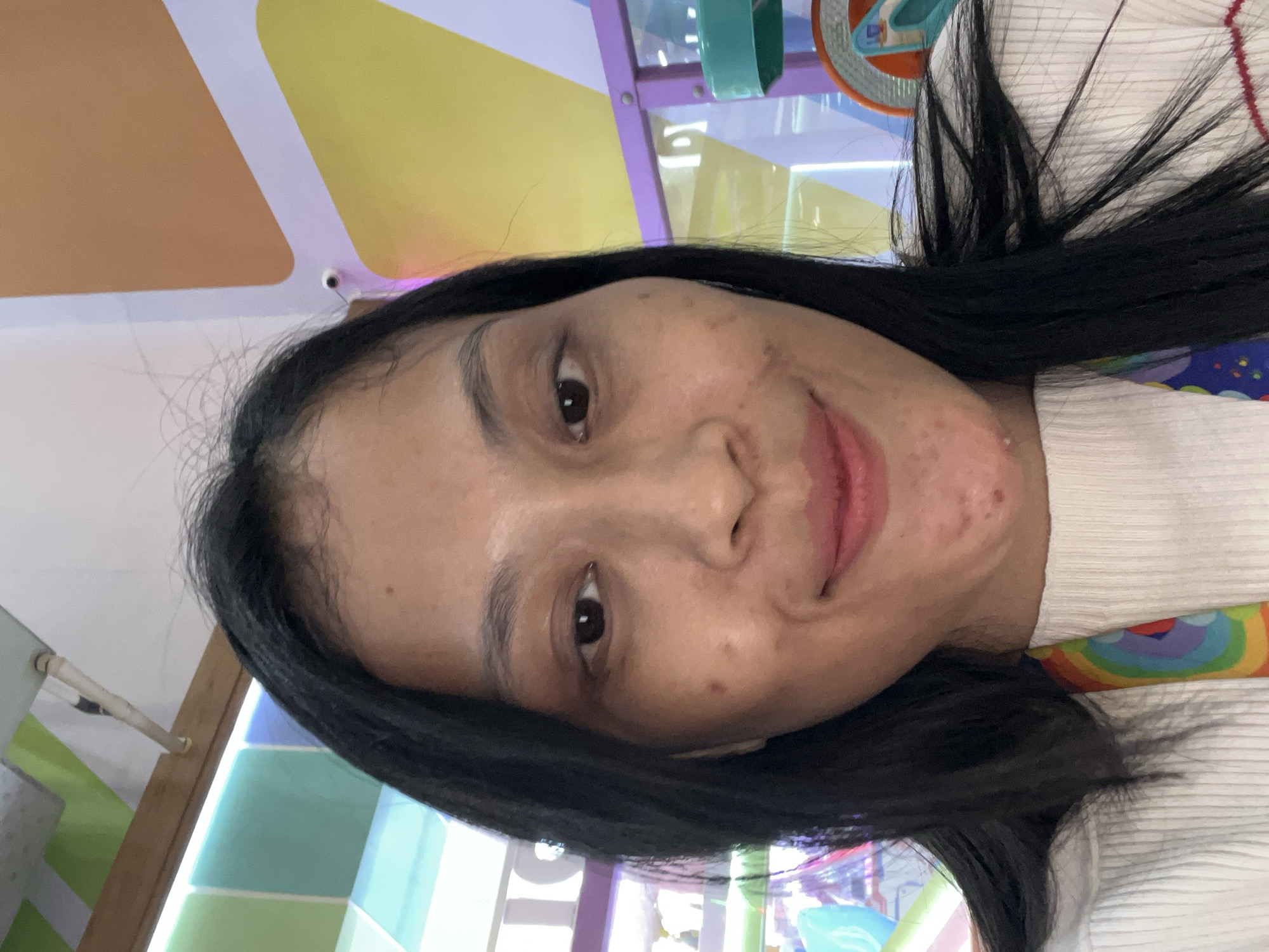 A medium shot, taken from a slightly low angle, captures a young Indonesian woman with long, dark hair framing her face. She is looking directly at the camera with a slight smile. The background is a colorful, abstract wall with geometric shapes in yellow, orange, and purple. Above her head, part of a wooden beam and what appears to be a ventilation system are visible. The woman is wearing a white, ribbed sweater, and a rainbow-colored lanyard is visible around her neck. The overall impression is that of a casual, possibly indoor, setting. The specific location of Kecamatan Pati, Indonesia, is not directly discernible from the visual cues in the image itself.