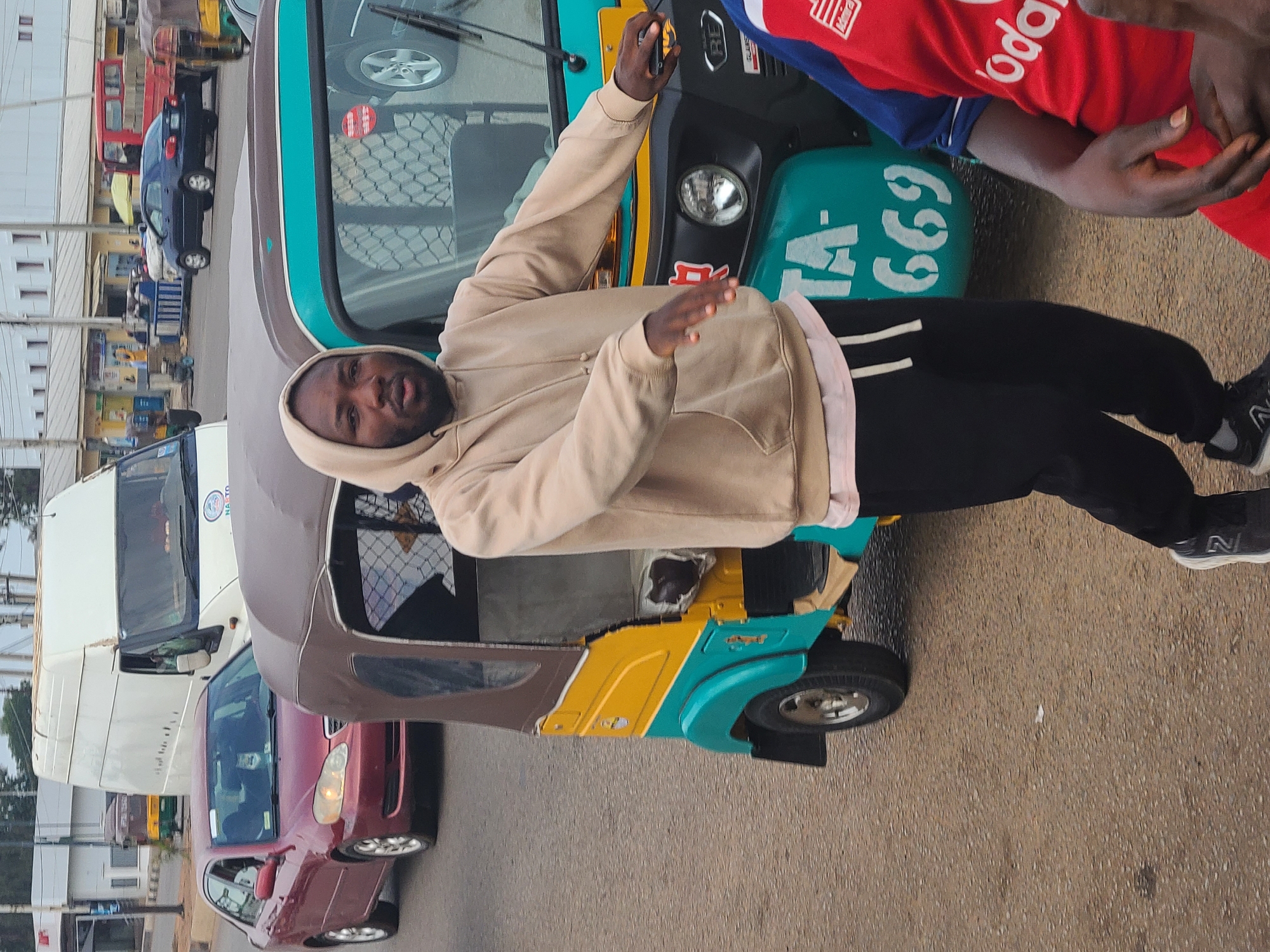 A man wearing a tan hoodie and black sweatpants is standing in front of a turquoise and yellow Bajaj Qute (also known as a "Keke Napep" or "Maruwa") in an urban environment. The vehicle has the number "TA-669" on its front. Another person, wearing a red Vodafone-branded jersey, is standing near the man. Cars and buildings are visible in the background. The location appears to be a busy street.
