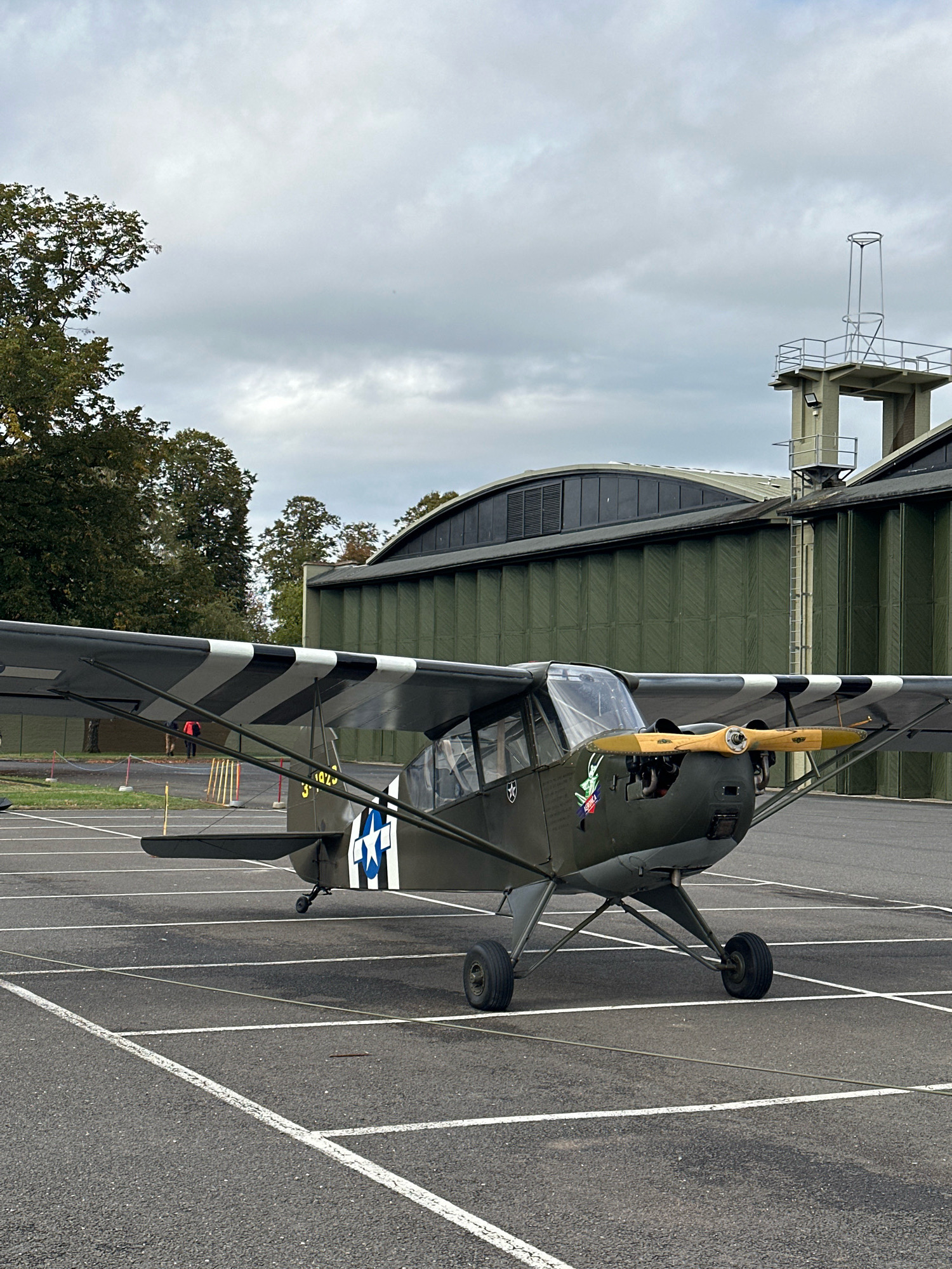 This image depicts a military-style airplane parked on an asphalt surface marked with white parking lines. The airplane, a dark green, single-engine, high-wing aircraft with a wooden propeller, features black and white stripes on its wings and a blue star insignia. In the background, several large, olive-green hangars with arched roofs are visible, along with a control tower structure. Trees with autumn foliage are present to the left of the hangars. Two individuals, one in an orange top and the other in darker clothing, are standing in the distance to the left of the aircraft. The sky is overcast with gray clouds. The scene appears to be set at an airfield in South Cambridgeshire, United Kingdom, likely Duxford Airfield, given the presence of vintage aircraft and hangars typical of the Imperial War Museum collection housed there.