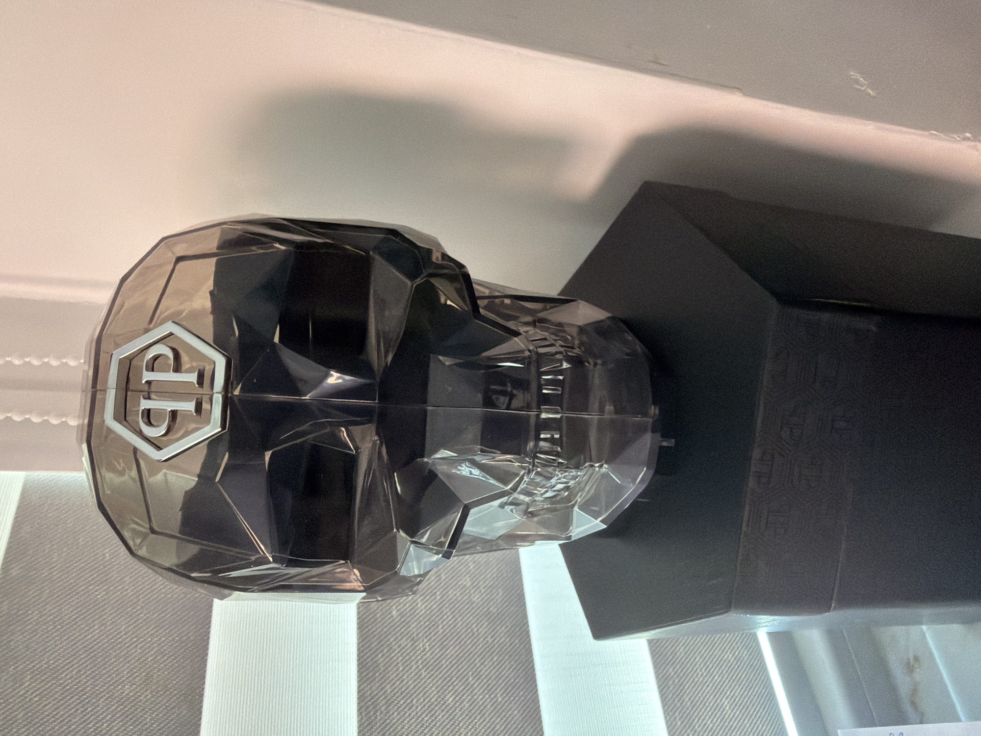 The image depicts an indoor scene in Sofia, Bulgaria, centered on a dark, translucent, faceted skull-shaped object. This object features a polished silver hexagonal emblem with the letters "PP" centrally positioned on its forehead. The skull rests on a dark, hexagonal pedestal that exhibits a subtle, embossed repeating pattern along its visible side. To the left, a window with horizontal blinds, striped with alternating light and dark bands, is partially visible, allowing diffused light into the space. To the right, a plain, light-colored wall is present, displaying minor surface imperfections and a soft shadow. No human subjects or dynamic actions are depicted.