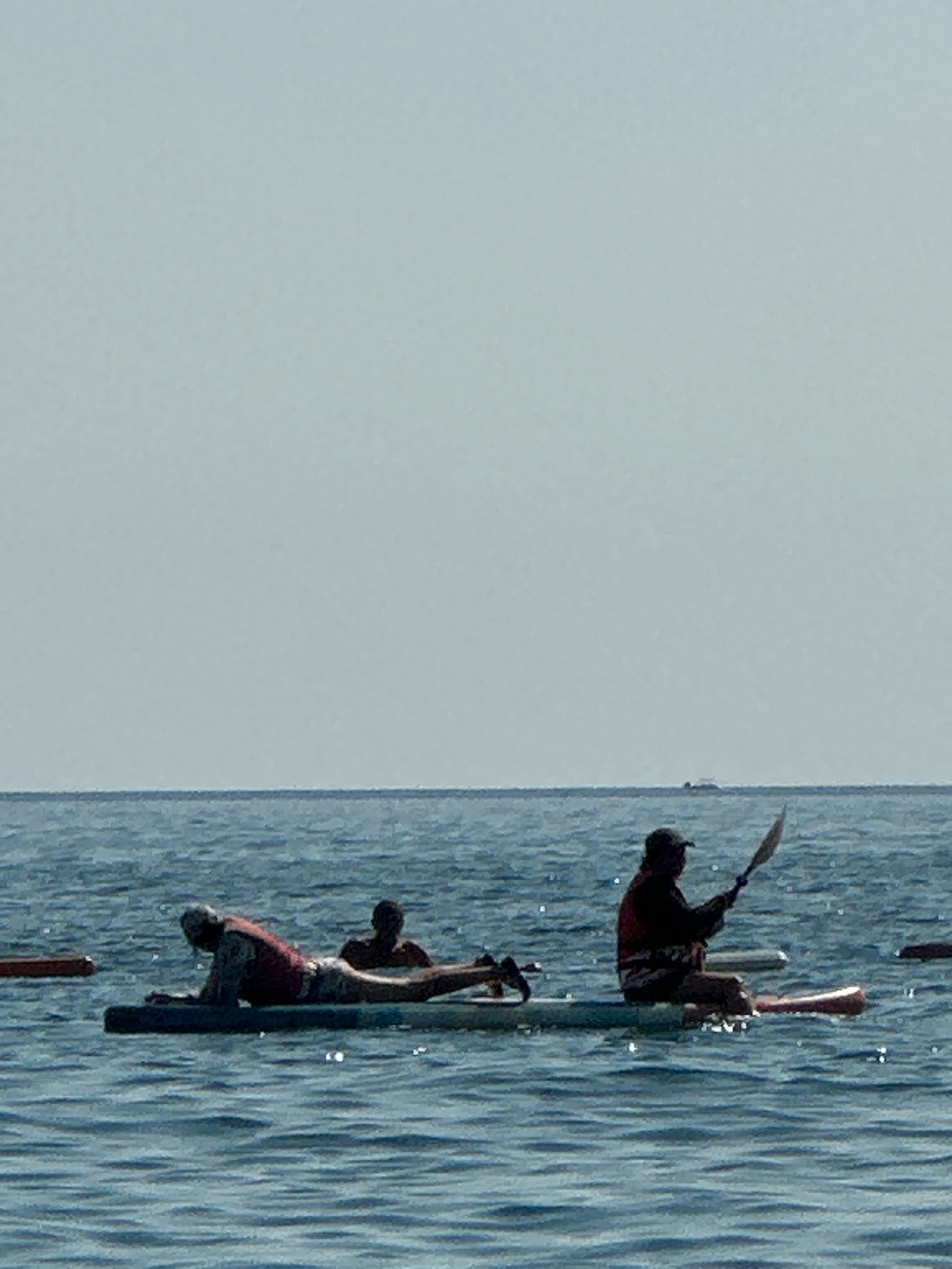 The image depicts a serene scene on the water, likely the Adriatic Sea, with the hazy silhouette of distant landmasses on the horizon. The sky is a pale, washed-out blue, suggesting a bright but potentially overcast or hazy day. The water is a muted blue-gray, with gentle ripples indicating calm conditions.
In the foreground, three individuals are engaged in water activities on paddleboards. On the left, a person is lying prone on a paddleboard, wearing a life vest and what appears to be shorts. Their body is angled towards the viewer. To their right, another person is partially submerged, with only their head and shoulders visible, also appearing to be on a paddleboard. Further to the right, a third individual is seated on a paddleboard, facing left and holding a paddle. They are wearing a life vest and shorts.
Several paddleboards are visible, some with individuals on them, and others appearing to be unattended. The overall impression is one of leisurely water recreation on a calm day. The location is described as Boreti, Montenegro, suggesting this scene takes place off the coast of this region.