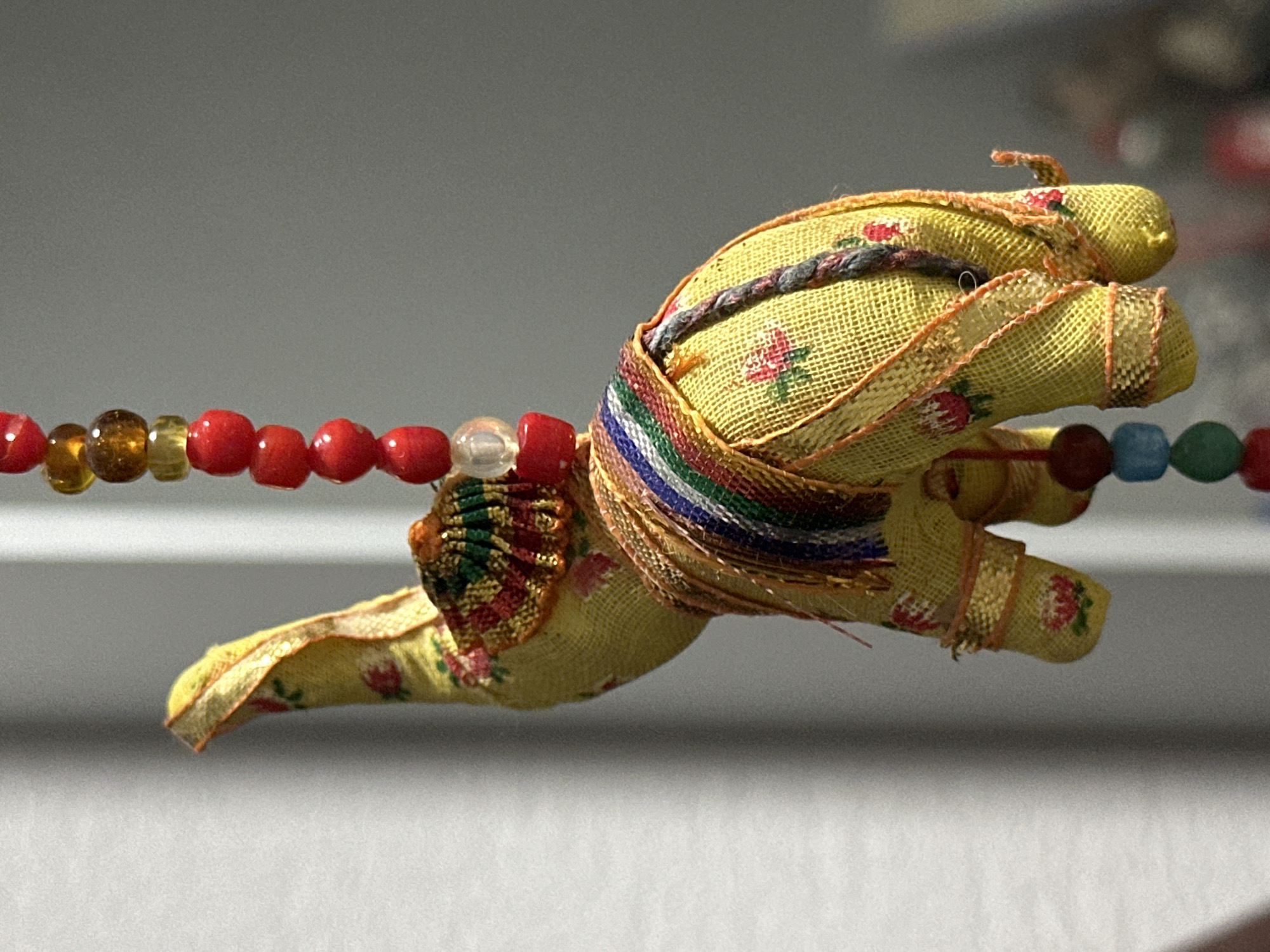 A close-up, eye-level shot shows a small, yellow fabric elephant figurine with a floral pattern hanging against a blurred background. The elephant is decorated with colorful trim and beaded elements. Its trunk is raised, and its legs are positioned as if it were standing. A strand of red, yellow, and clear beads hangs from the top of the elephant, continuing downwards. The lighting suggests the object is indoors, illuminated by natural light casting shadows. The background consists of indistinct vertical lines on the left, likely a window blind, and a plain grey surface on the right. The location is not specified in the image.