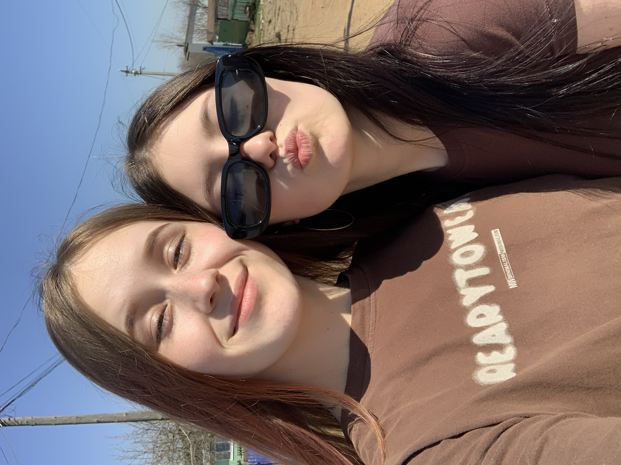Two young women are posing close together for a selfie outdoors. The woman in the foreground has light brown hair with pinkish highlights, light skin, and is smiling with her eyes mostly closed. The woman in the background has long, dark brown hair and is wearing large, square black sunglasses and pursing her lips in a kissing expression. They are both wearing brown shirts. The shirt of the woman in the foreground has white text that reads "DEADTOW". The background appears to be a street or rural area, with a clear blue sky visible in the upper left. Based on the location provided, the photo was taken in Фоки, Russia.