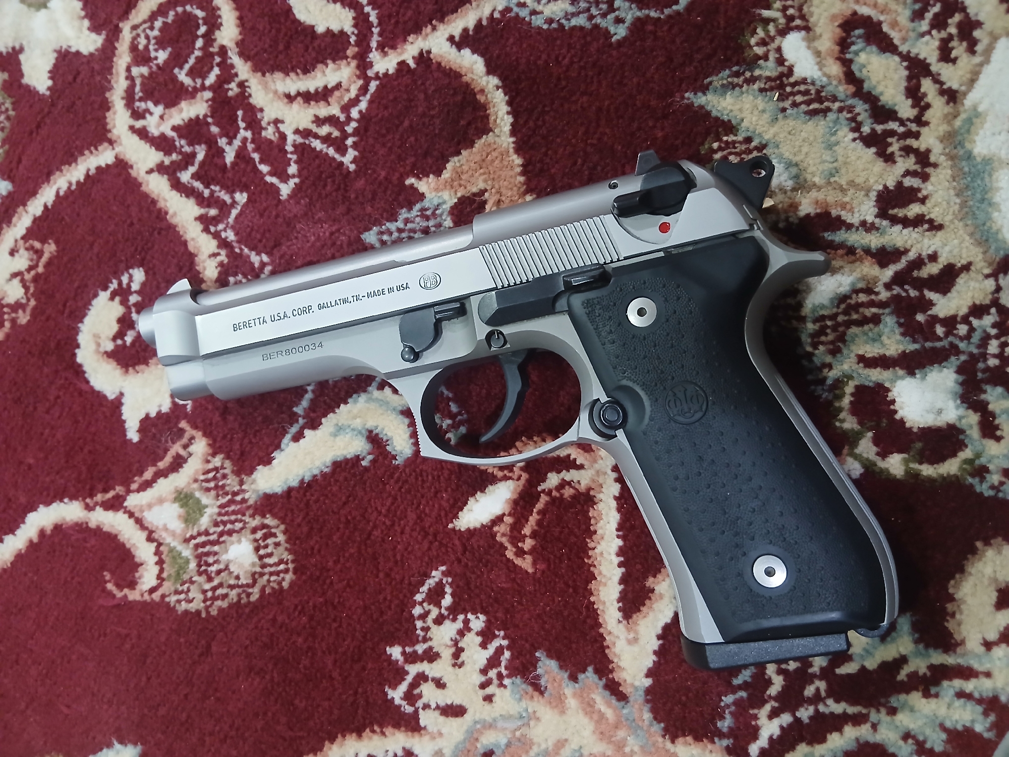 A Beretta 92FS Inox pistol lies on a patterned carpet. The pistol is silver with black grips, oriented diagonally across the frame. The text "BERETTA U.S.A. CORP. GALLATIN, TN - MADE IN USA" is visible on the slide, as is the serial number "BER800034". The carpet has a complex design in shades of red, white, and brown. The environment is assumed to be indoors. There is no indication of the location being Quetta City Tehsil, Pakistan. No people, actions, or interactions are visible.