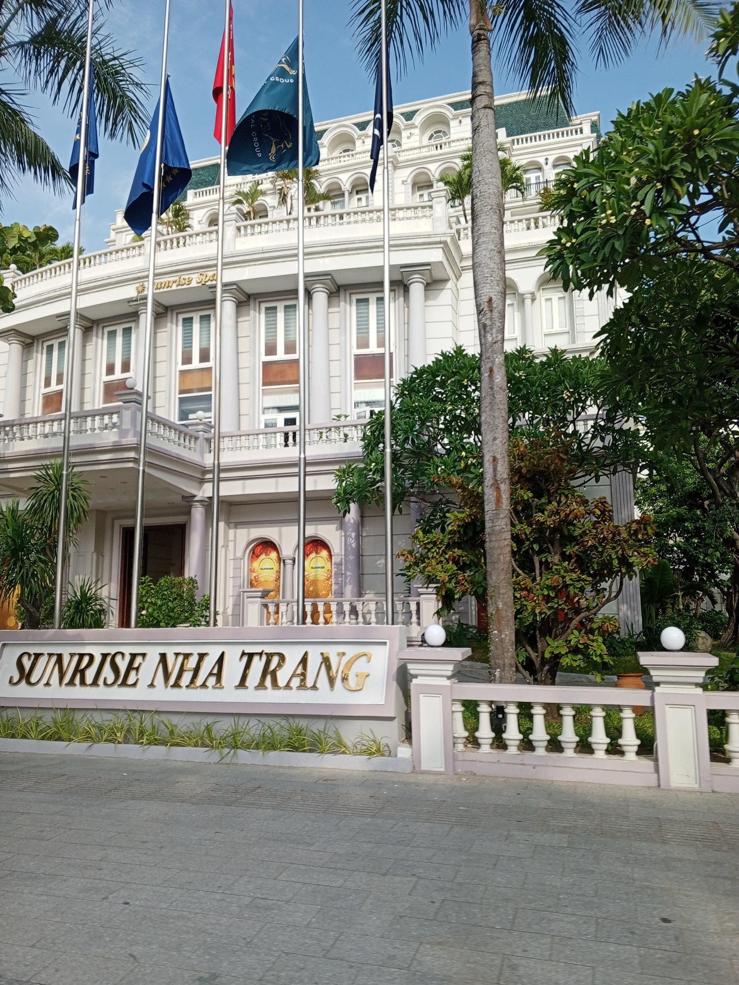 The image shows a hotel entrance with a large sign reading "SUNRISE NHA TRANG". The hotel building is white with ornate architectural details, including columns and balconies. There are flags flying in front of the building. Palm trees and other lush greenery surround the entrance. The sky is clear and blue. The pavement in front of the sign is made of grey square tiles. This scene is typical of a tropical resort or hotel in Nha Trang, Vietnam.