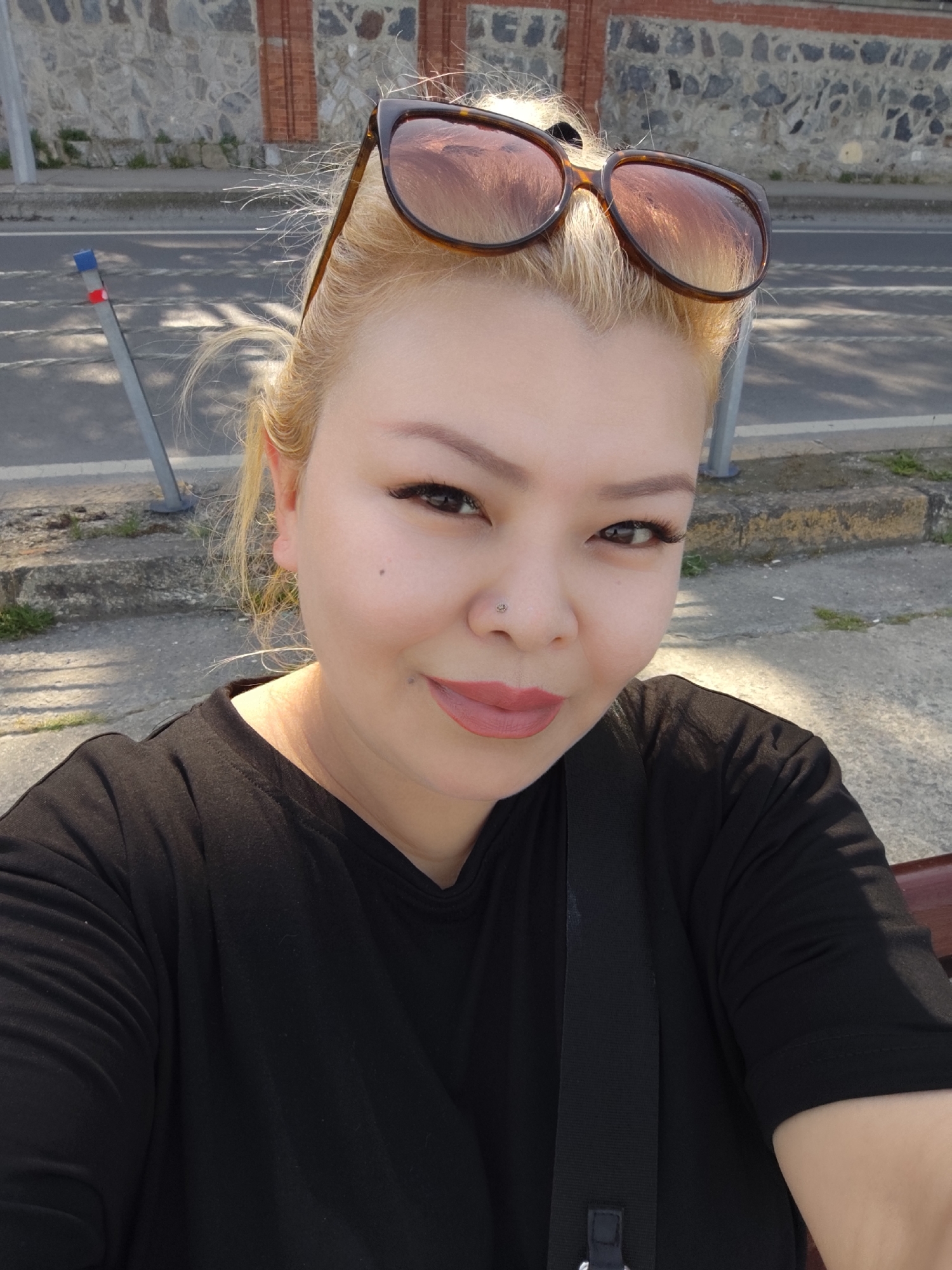 A woman with blonde hair and brown sunglasses on her head is taking a selfie outdoors. She has a nose piercing and is wearing a black t-shirt. In the background, there is a road, a sidewalk, and a stone wall with brick accents. The location is Sarıyer, Turkey, based on recognizable architectural elements.