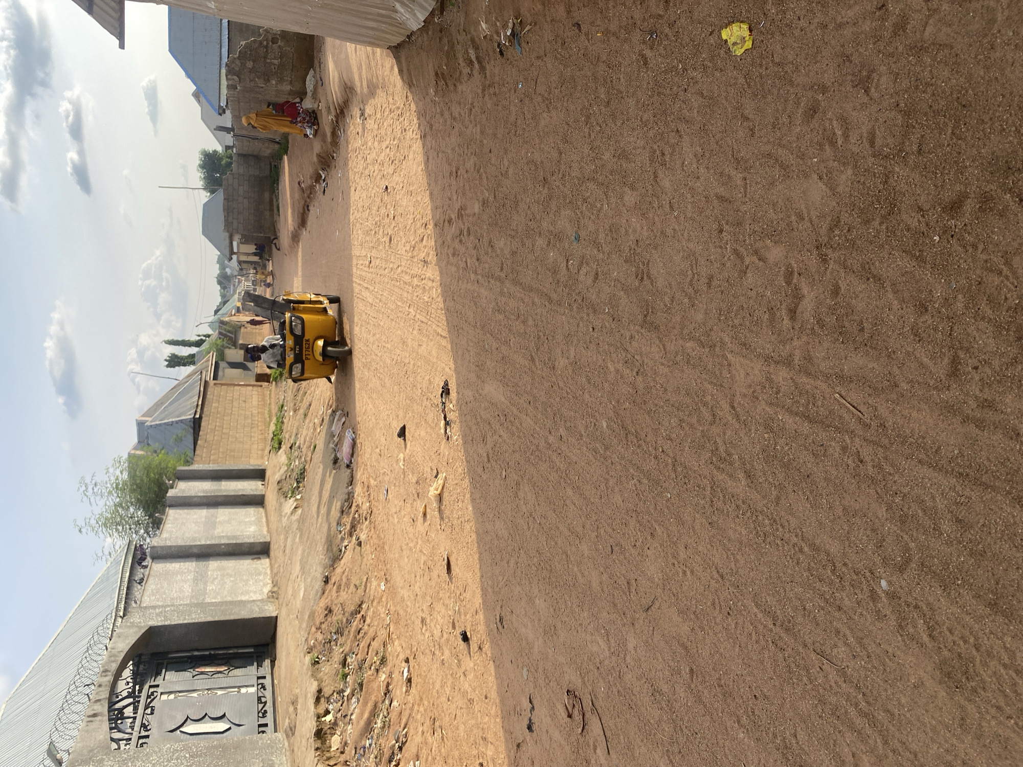 This image depicts a dusty, unpaved street in a populated area. A bright yellow tricycle, a common mode of transport in some regions, is prominently featured in the center of the frame, carrying at least two passengers. The street is lined with buildings constructed from various materials, including cinder blocks and corrugated metal, suggesting a mix of residential and possibly commercial structures. Some of the buildings appear to be in disrepair, with debris scattered on the ground.
In the background, a few individuals are visible, some walking along the street. One person is wearing a yellow head covering and a red garment, while others are dressed in more muted clothing. The sky is partly cloudy, with a few distinct cloud formations visible. The overall impression is of a warm, sunny day in a place where infrastructure may be developing. The lighting suggests it is likely late morning or early afternoon. The ground is covered in fine, dry dirt, with some evidence of tire tracks from vehicles. There is also a small amount of litter present on the side of the road. The scene appears to be a typical day in a town or village, with people going about their daily activities. The image is oriented vertically, which may affect the perspective of the scene. There is some visible text on the tricycle, specifically the license plate which reads "TVSPY 752K".