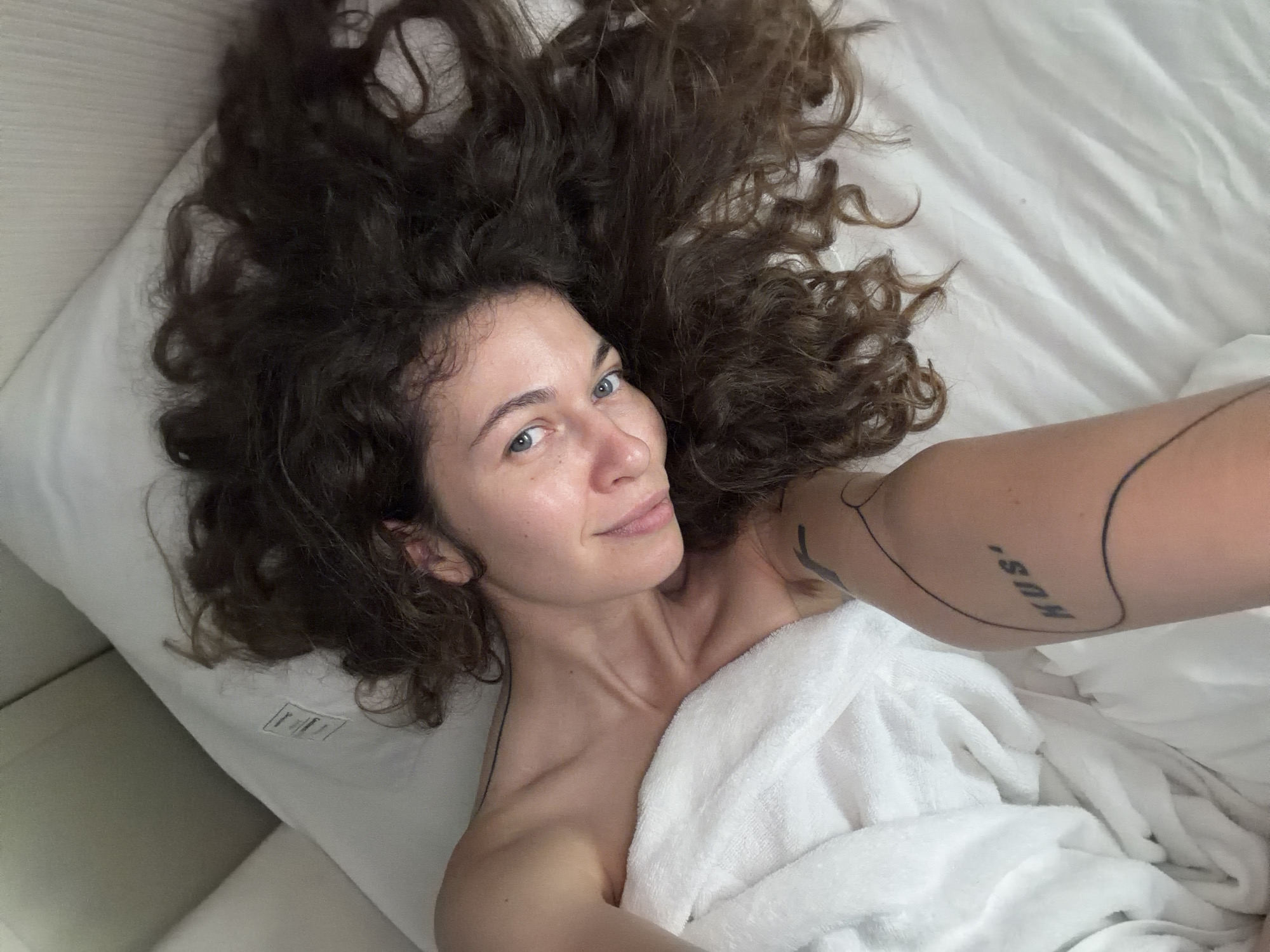 An overhead selfie captures a woman with voluminous, dark brown curly hair spread out across crisp white bed sheets and a pillow. She is lying on her back, looking directly at the camera with clear, light blue eyes and a subtle, relaxed smile. Her bare shoulders and chest are partially visible, covered by a soft white towel wrapped around her.
Her left arm, extended to take the photo, reveals several black line tattoos, including abstract lines and the distinct word "MUS'" on her forearm. The setting appears to be a bright room, likely a bedroom or hotel room in Hanoi, Vietnam, as suggested by the provided context. The natural light indicates it is daytime. A small white tag with partially legible text, possibly "I'm | FIT," is visible on the pillow near her head. The overall mood is one of serenity and comfort, suggesting a moment of rest or shortly after waking. Her complexion is natural and clear, with minimal or no makeup.