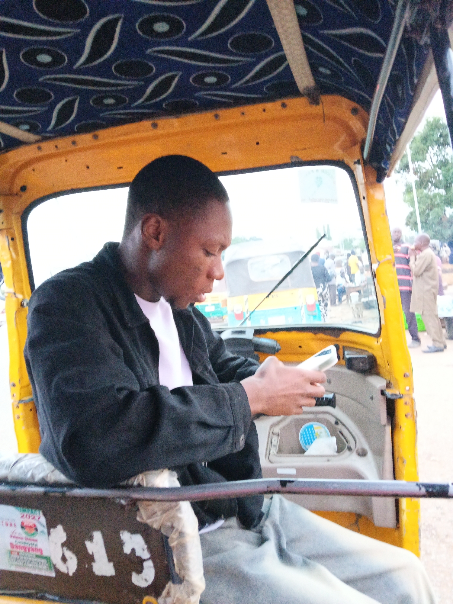 A young Black male wearing a black jacket over a white t-shirt sits in the driver's seat of a yellow auto-rickshaw. He is looking down and appears to be operating a white cell phone in his hands. The auto-rickshaw's interior has a patterned blue fabric lining the ceiling. Outside the vehicle, blurred figures and trees are visible. The auto-rickshaw has the number "613" painted on its side.
