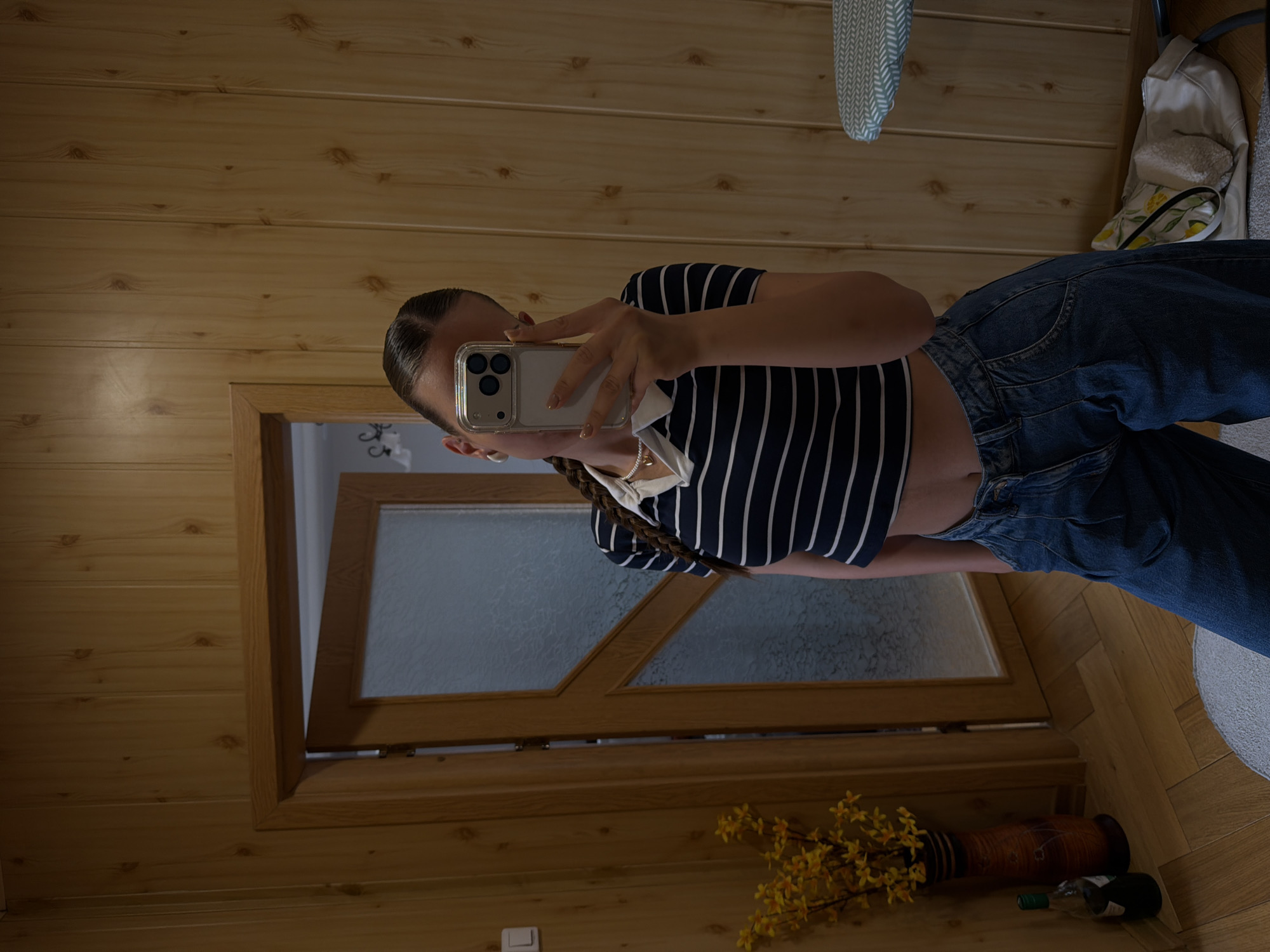A young woman is taking a selfie in what appears to be a wood-paneled room. She is wearing a navy and white striped crop top and blue jeans. Her hair is styled in a braid. A wooden door is visible behind her. On the floor near the door, there is a vase with yellow flowers and an empty wine bottle. The location is not discernible as Buhovo, Bulgaria based solely on visual data. The scene suggests a domestic setting rather than indicating a specific city.