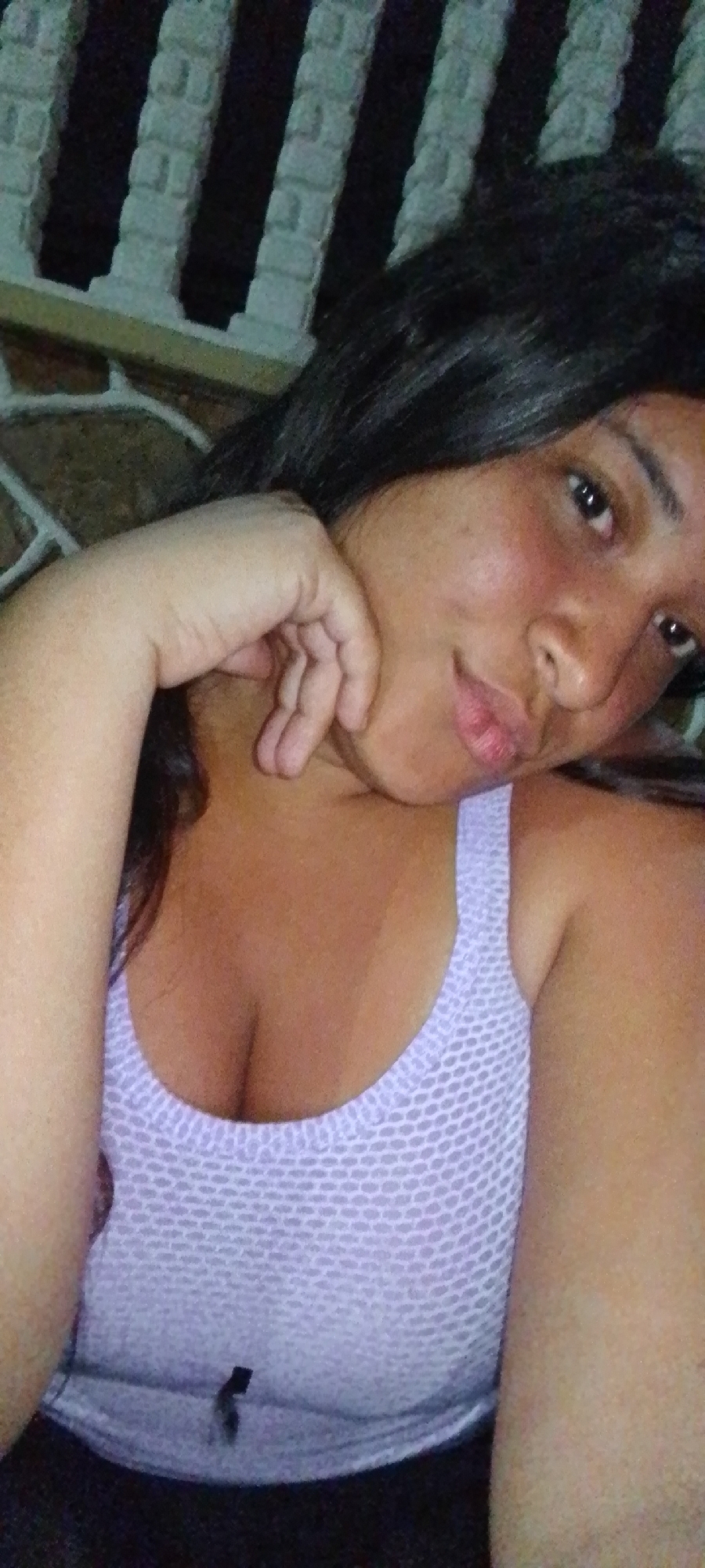 A woman is in the foreground, posing for a selfie. She is wearing a light purple tank top. Her black hair is loose. Her left hand is near her face. Behind her is a textured wall and a white railing. Based on the prompt, the location is the city of La Cabrera, Venezuela.