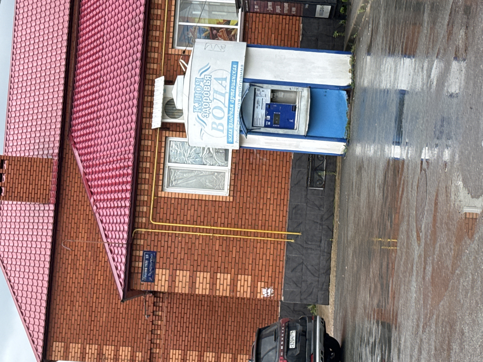 The image depicts an outdoor scene, likely in Kazan, Russia, with a focus on a water dispensing machine. The weather appears to be rainy, as the ground is wet and reflective, and the overall lighting suggests an overcast or late afternoon/early morning time.
The main subject is a blue and white water vending machine mounted on a brick building. The machine has a prominent sign with text in Russian that reads "КЛЮЧ ЗДОРОВЬЯ ВОДА" (Klyuch Zdorovya Voda), meaning "Key of Health Water." Below this, smaller text states "Артезианская" (Artezianckaya), indicating artesian water, and "Республиканская программа" (Respublikanskaya programma), suggesting a republican program. The machine itself has a blue front panel with a slot for payment and a dispenser area.
The building features a textured red brick facade and a distinctive pink tiled roof with a wave-like pattern. There are windows on the building, one of which is covered with a translucent material. A yellow pipe runs horizontally along the brick wall. To the left of the water machine, an air conditioning unit is visible.
In the foreground, a dark-colored SUV is partially visible, with its rear end facing the camera. The license plate is partially obscured but shows Cyrillic characters and numbers. The wet asphalt suggests recent rain.
A street sign is mounted on the brick wall, also displaying Cyrillic text, which appears to be the name of a street and possibly a building number. The overall impression is one of a functional urban or suburban setting, with the water dispenser being a key feature of the scene. There are no people clearly visible in the image.