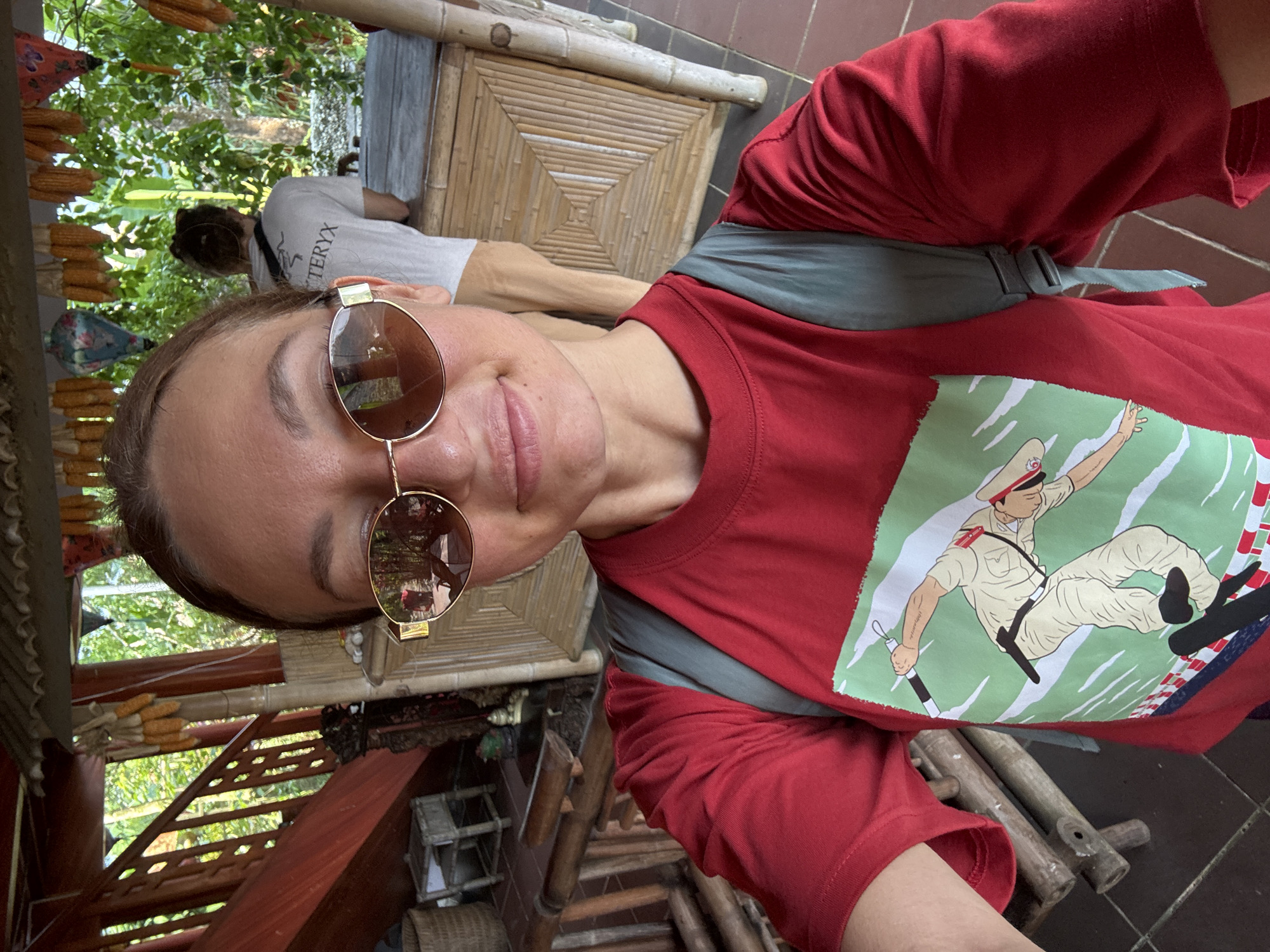 This selfie image was captured in what appears to be an indoor or semi-outdoor establishment in Thông Nguyên Commune, Vietnam, during daylight hours. The primary subject is a person with short brown hair, wearing a red t-shirt, round gold-rimmed sunglasses, and a gray sling bag or backpack. They are seen with a slight smile, suggesting contentment.
The red t-shirt features a prominent graphic depicting a uniformed figure, possibly a police officer or soldier, in a dynamic pose. This figure is shown holding a baton or stick, against a green background with white stripes or waves, potentially illustrating an action like skateboarding or surfing.
In the background, another individual is visible from behind, wearing a white t-shirt that clearly displays the "ARC'TERYX" logo and light-colored pants.
The setting incorporates numerous rustic and traditional elements. Decorative strings of dried corn cobs hang from an overhead structure, and the furniture, including a table, appears to be crafted from bamboo. Wooden beams, stairs, and other structures are also present, alongside woven baskets. Beyond the immediate structures, lush green foliage is visible, indicating an outdoor or natural environment surrounding the establishment. The overall atmosphere points to a rural or traditional Vietnamese locale.