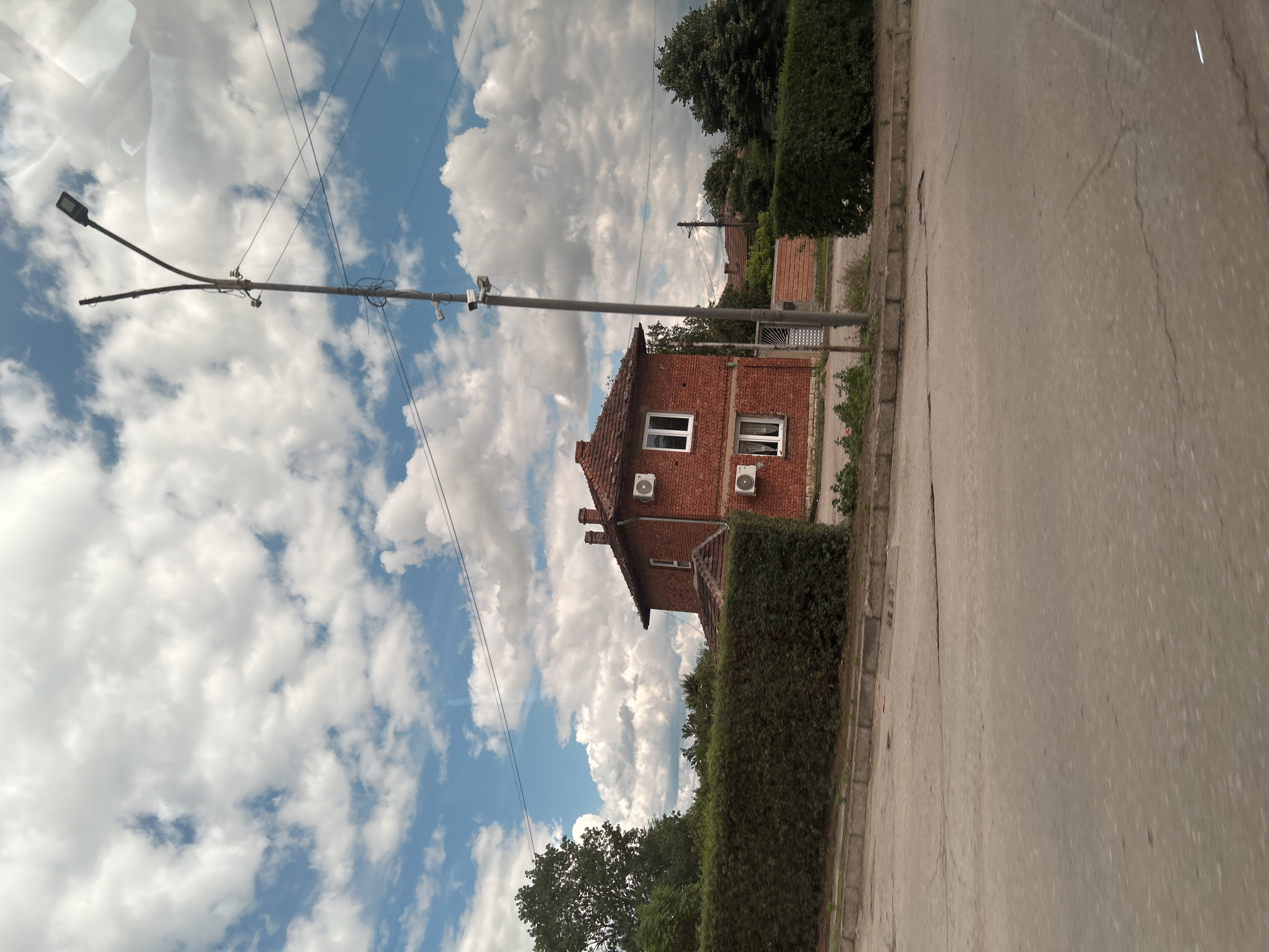 The image captures a street view in an urban environment under a cloudy sky. A two-story brick house with a red tile roof is the central subject, featuring two visible air conditioning units mounted on its exterior. The house is partially obscured by a dense, well-maintained green hedge in the foreground. A tall lamppost with a modern LED fixture and what appear to be security cameras is positioned to the right of the house. Power lines are strung across the sky, connecting to the lamppost and extending out of frame. The street in the foreground shows signs of wear, with cracks and uneven patches. Behind the hedge and house, more trees and buildings are visible, suggesting a residential or mixed-use area. The overall impression is that of a moderately developed urban setting on a partly cloudy day.