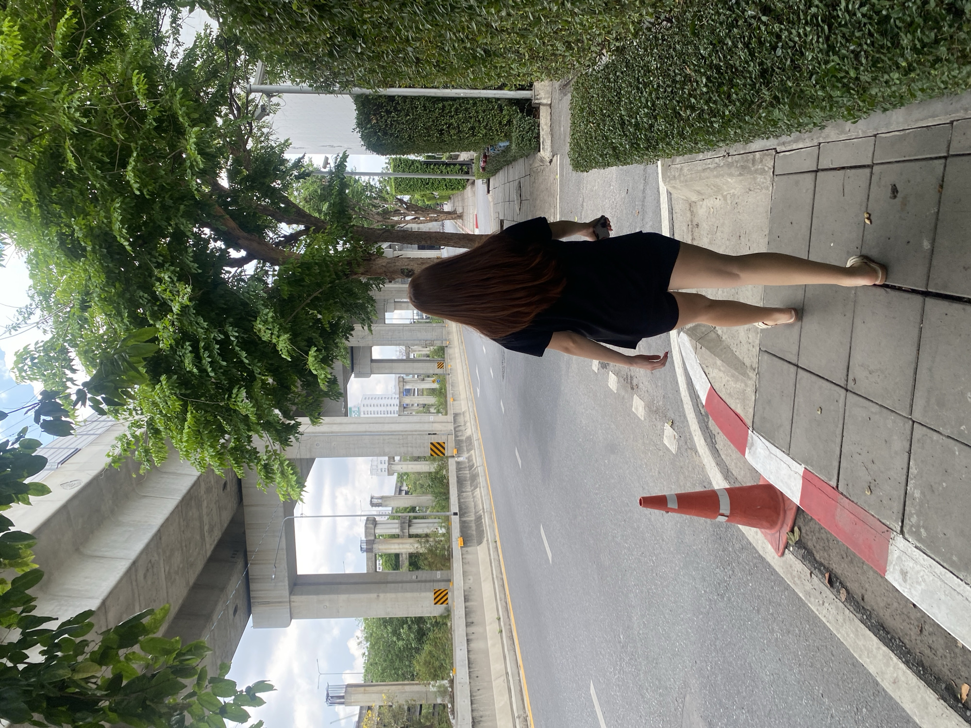 The image depicts a woman walking on a sidewalk in Bangkok, Thailand. She has long, brown hair and is wearing a black dress and light-colored sandals. The sidewalk borders a road with an orange traffic cone at the edge. An elevated highway structure is visible in the background, with trees and greenery lining the roadside and sidewalk. The sky is overcast.