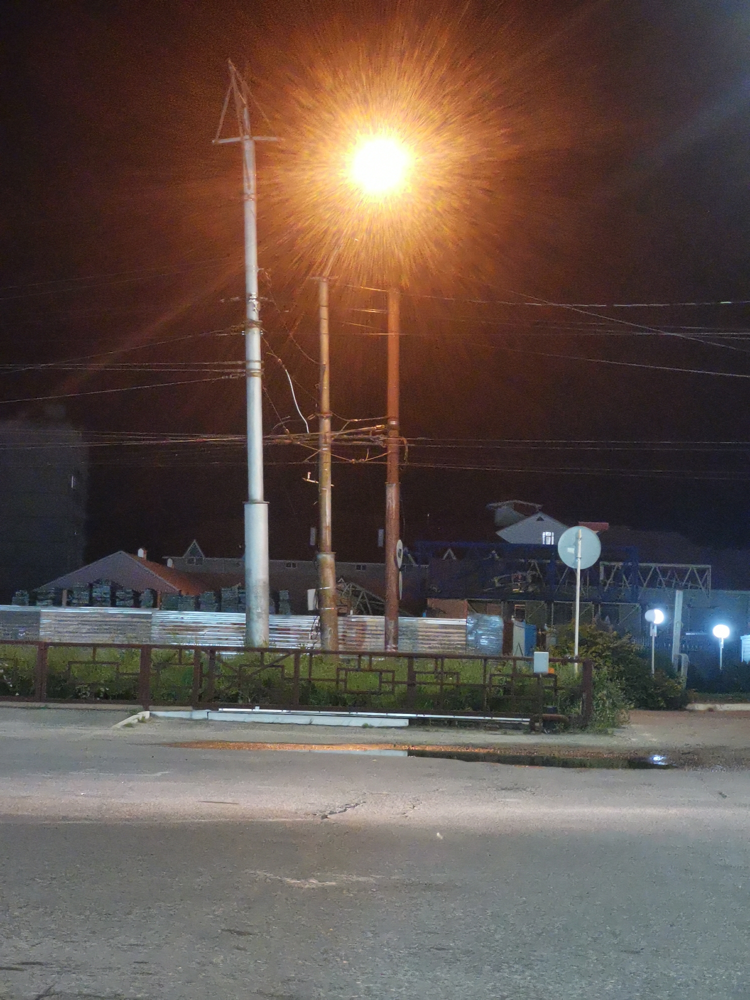 A night-time street view captures a bright, starburst-like street light illuminating the scene. Two prominent utility poles stand tall, one white and the other weathered brown, supporting a network of overhead wires. In the background, a cluster of buildings with dark roofs and some visible windows suggests a residential or commercial area. A rusted metal fence with a green, overgrown barrier runs horizontally across the mid-ground. The foreground features an asphalt road with some reflective puddles. A circular white road sign on a pole is visible to the right of the white utility pole, and a second, smaller white sign is seen behind it. The overall ambiance is that of a quiet, dimly lit urban environment at night. The scene is consistent with a street in Cherkessk, Russia, given the architectural style and common utility infrastructure. There are no people or animals present in the image.
