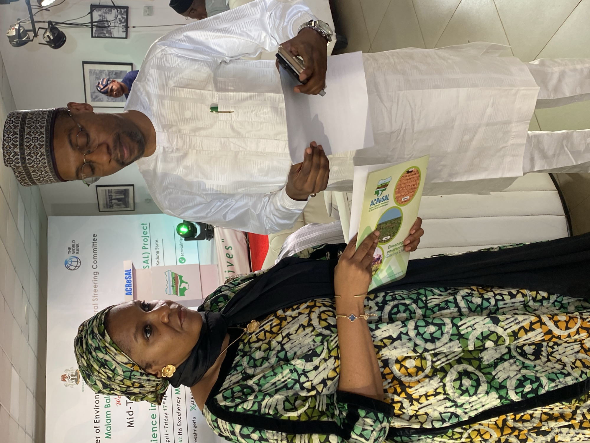 The image shows a man and a woman standing in front of a banner. The man is wearing a white outfit and a hat, and he is holding a white paper and a cell phone. The woman is wearing a patterned green dress and a headwrap, and she is holding a brochure. The banner behind them has the words "ACRESAL", "Kaduna State", "The World Bank", "ral Steering Committee", and "Saving Live". The text visible also includes "Minister of Environment", "Malam Bala", "Mid-Term Review", and "Kwarau, Nigeria" can be seen on the poster, indicating the location and setting of the picture.