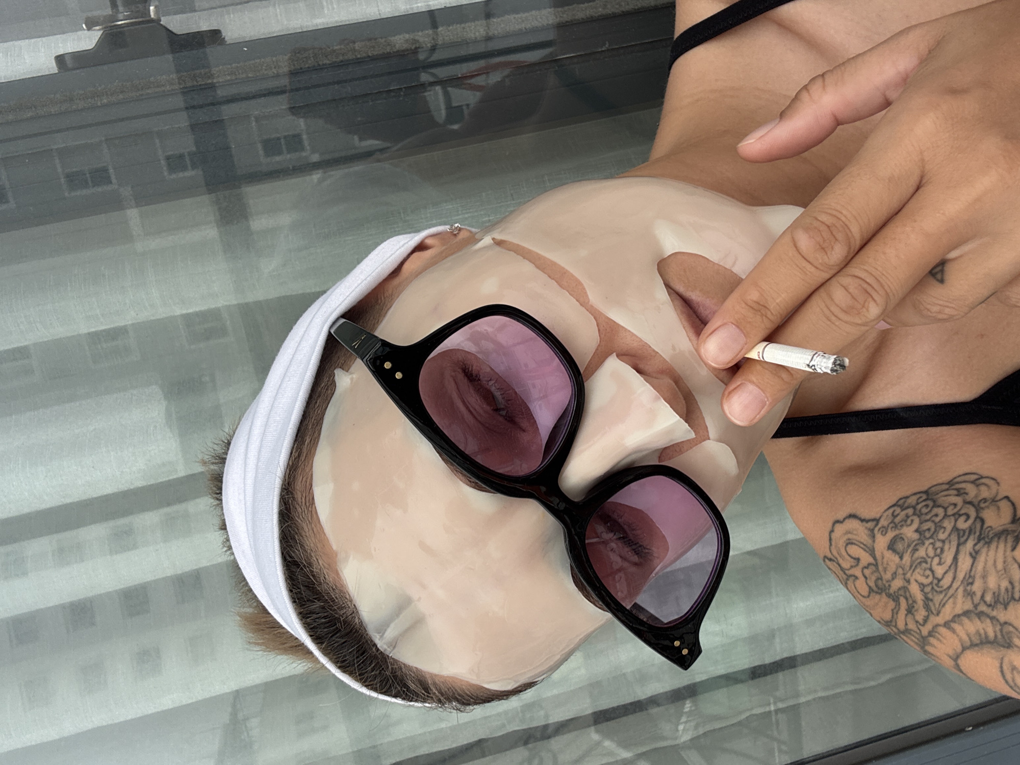 A person with a white headband and a sheet mask on their face is holding a lit cigarette between their fingers. They are wearing black cat-eye sunglasses with pink lenses, and a portion of a tattoo is visible on their left shoulder. The person is wearing a black top. The background consists of a window with sheer curtains, revealing a building with multiple windows. The setting appears to be indoors, possibly an apartment or hotel room, with a view of a cityscape. There are no direct indications of the location being specifically in Nha Trang, Vietnam.