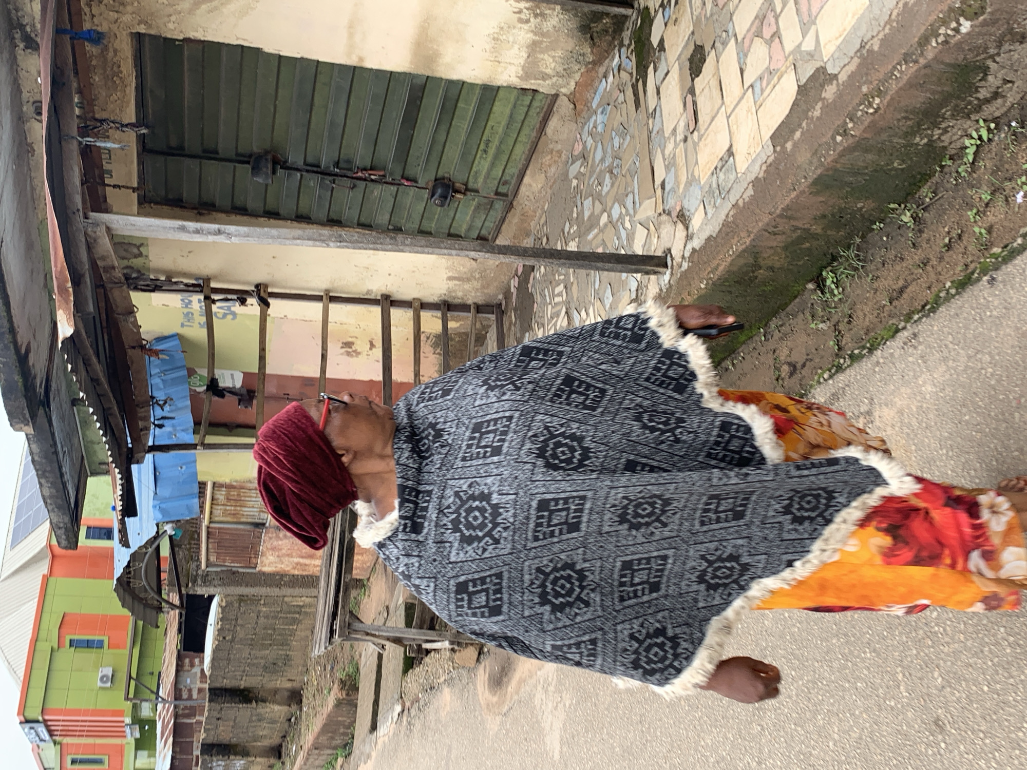 A woman in a dark patterned poncho and a red headscarf walks on a paved street in Jos, Nigeria. She is wearing glasses and a colorful, floral-patterned dress or skirt underneath the poncho. The street appears to be lined with buildings. The building on the right has a green metal shuttered door and a beige, weathered wall. The ground is paved with irregular tiles in varying shades of tan and gray. A concrete drain runs along the side of the street. In the background, a brightly colored building with green and orange sections and a white roof is visible, along with other structures that appear to be shops or houses with corrugated metal roofs. The sky is overcast.