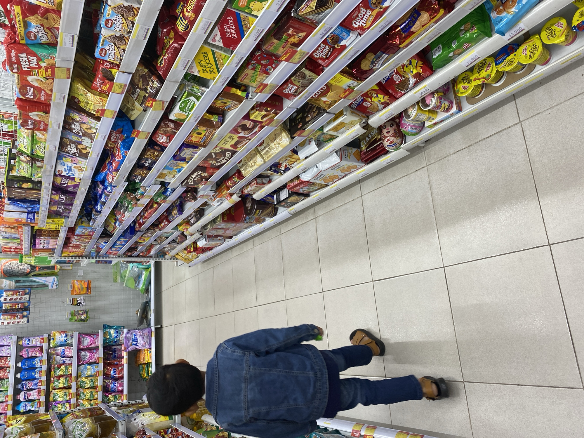 A young child, seen from the back, walks down an aisle in a supermarket. The child is wearing a denim jacket, dark pants, and brown sandals. The aisle is lined with shelves stocked with various food products, primarily snacks and confectionery. The flooring is tiled in a light, neutral color. The lighting appears to be artificial, typical of an indoor retail environment.
