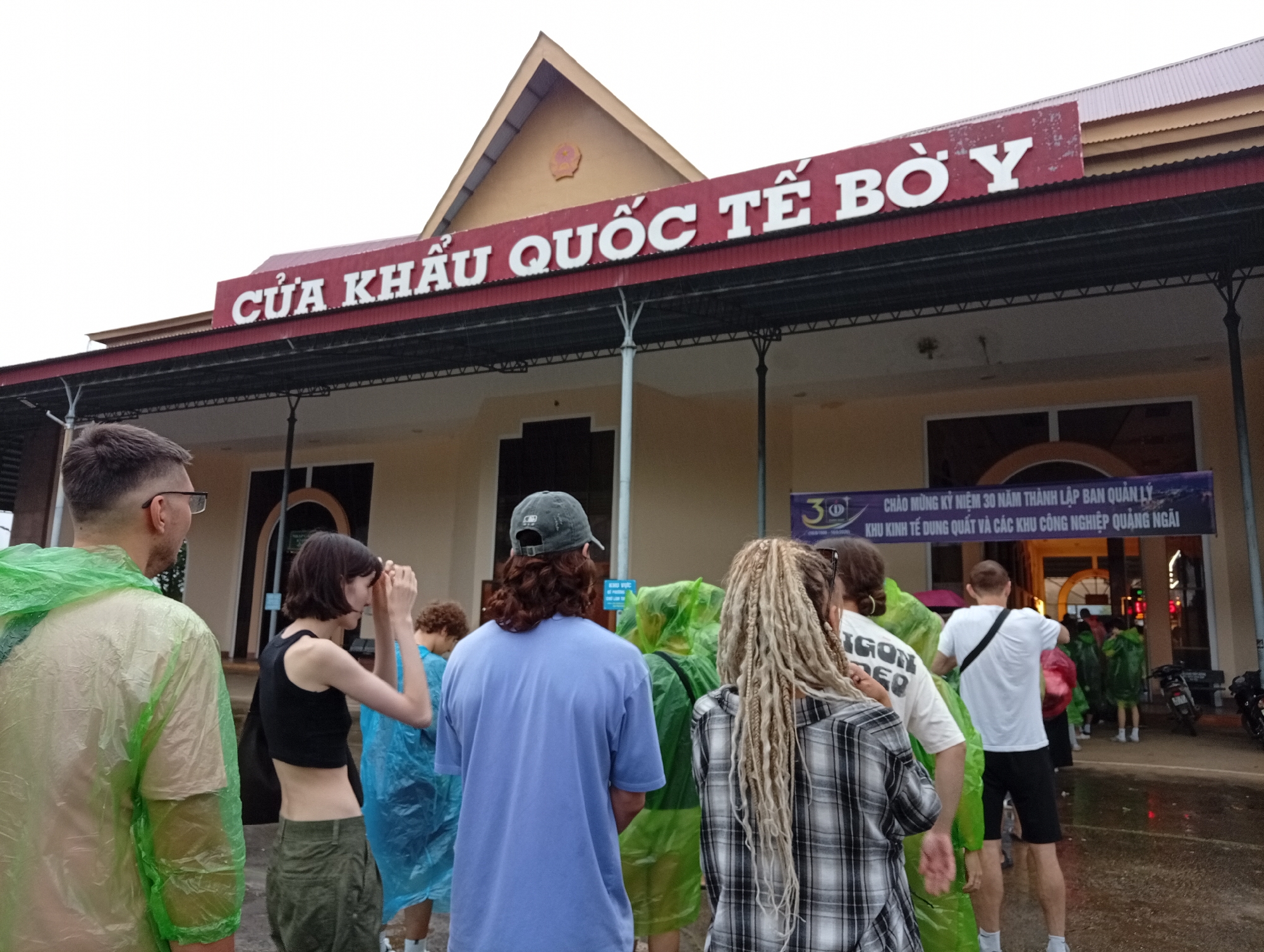 In the city of Bo Y, Vietnam, a group of people are gathered outside a building with a large red sign above the entrance that reads "CỬA KHẨU QUỐC TẾ BỜ Y". The building appears to be a border crossing or international gateway. Most of the people are wearing transparent or light-colored raincoats, suggesting it might be raining or has recently rained, as indicated by the wet ground.
In the foreground, from left to right, a man wearing a light green raincoat stands with his back to the camera. Next to him, a woman in a black crop top and green pants is looking forward and adjusting her sunglasses. Further back, a person in a blue raincoat is visible, and to their right, a person with blonde dreadlocks in a black and white plaid shirt is facing away. In the background, several other individuals are also seen wearing raincoats, some entering the building or standing near a banner. The banner, draped across the building, contains Vietnamese text, possibly celebrating an anniversary or event related to the border and industrial zones. Motorcycles are parked to the right of the scene. The overall atmosphere suggests a travel or transit point, possibly experiencing inclement weather.