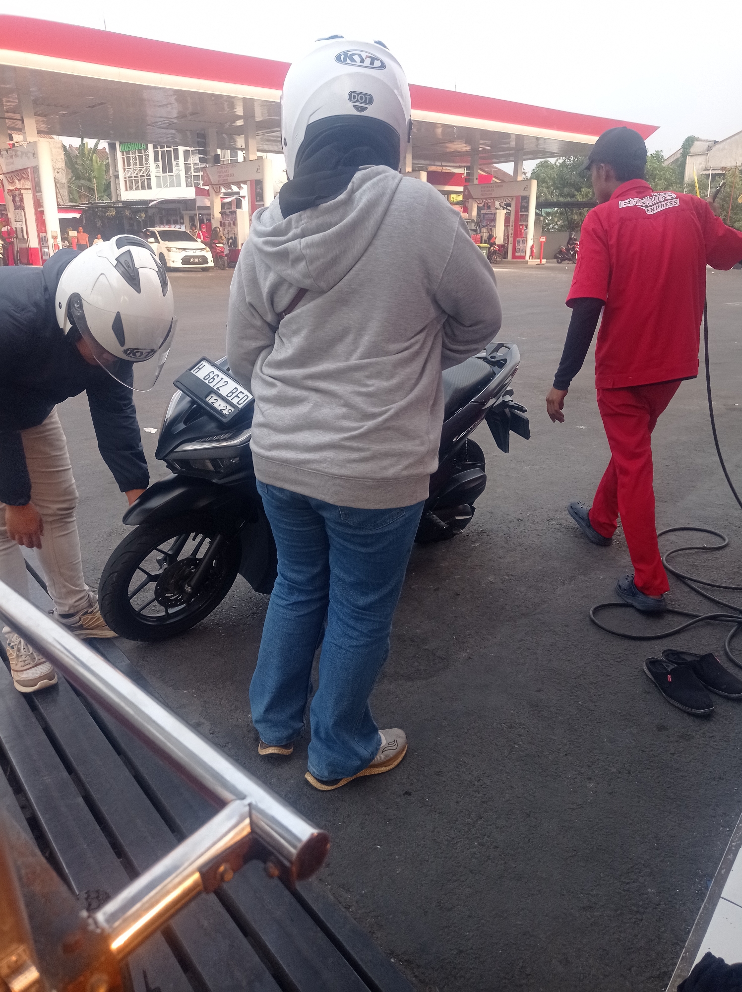 At a gas station in Bangunsari, Indonesia, two individuals wearing helmets are near a motorcycle. One, wearing a gray hoodie and blue jeans, stands behind the motorcycle. The other, wearing a black jacket and light-colored pants, is bending over to examine the front wheel of the motorcycle. A gas station attendant in a red uniform with a black undershirt is walking away from the motorcycle towards the right of the frame. The gas station canopy with red and white accents is visible in the background, along with a white car. The ground is paved asphalt, and to the left of the frame, a metallic handrail is present.