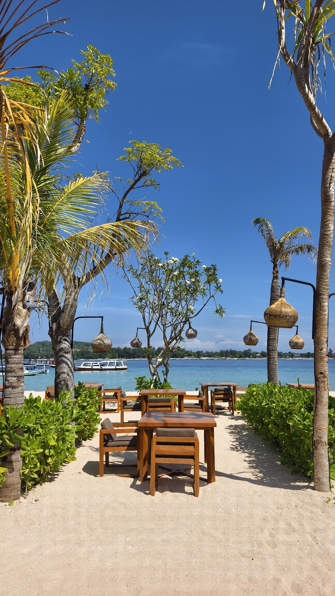 The image depicts an outdoor dining area on a beach, likely in an area known for tourism in Unknown, Indonesia. The scene features several wooden tables and chairs arranged on a sand path, bordered by lush green bushes. Palm trees are scattered throughout the area, some adorned with decorative woven lamps hanging from metal posts. In the background, the turquoise ocean stretches out under a clear blue sky. A boat is visible on the water, and a glimpse of land can be seen in the distance. The setting suggests a relaxed, tropical atmosphere, typical of a beachside restaurant or resort. There are no people visible in the image.