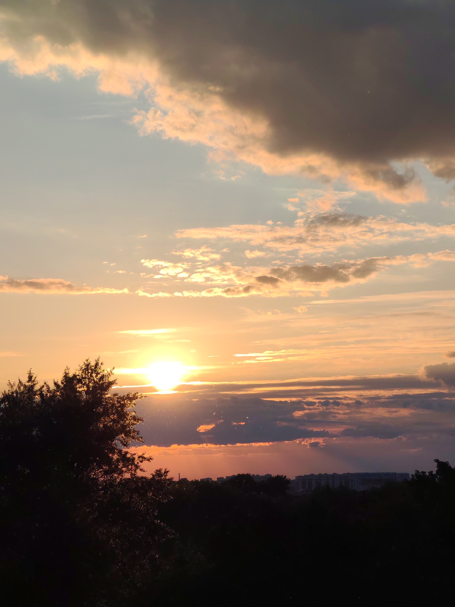 The media file presents a vibrant sunset over Podolsk, Russia, with the sun prominently featured on the left side of the horizon. The sky is a dominant subject, displaying a dramatic array of colors and cloud formations. Warm orange and golden hues emanate from the setting sun, illuminating surrounding clouds, which range from bright peach and light blue near the sun to darker grays and purples towards the upper right and lower right portions of the sky. Various cloud types, including scattered cumulus and layered stratocumulus, are visible.
The foreground is heavily silhouetted by the low light, featuring the dark, dense canopy of trees on the left and extending across the bottom of the frame. In the mid-ground, a hazy urban landscape with distant buildings and structures is visible in silhouette against the colorful horizon, suggesting a city or town extending into the distance. Specks of dust or light flare are noticeable in the air, particularly around the sun. The time of day is clearly dusk or sunset due to the low sun angle and the intense, warm coloration of the sky. There is no discernible activity or visible text in the image. The overall scene evokes a sense of serene beauty and the peaceful close of day.