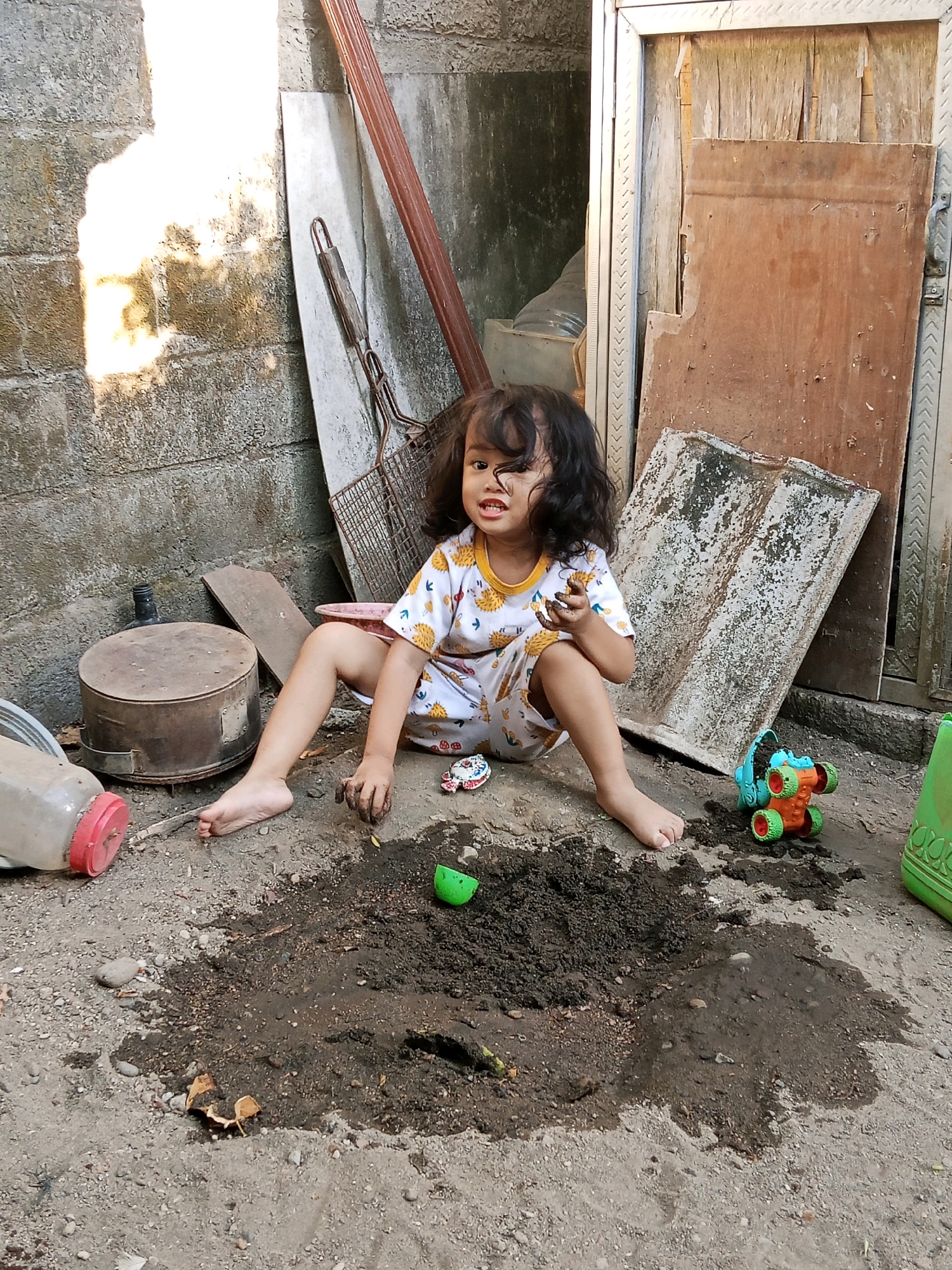 A young child with curly dark hair is playing with mud in the dirt yard of a residence located in Gaprang, Indonesia. The child, dressed in a white t-shirt with yellow trim and a pattern of hedgehog-like creatures, is seated with legs spread, their hands covered in mud as they interact with the soil. A small green plastic bowl is near the child, and a colorful toy truck with orange and green wheels sits to the right of the child. In the background, a rough concrete wall is visible, with sunlight casting a bright shadow on it. Lean against the wall are various items, including a metal grid with a wooden handle, a large round metal pot with a lid, and a piece of worn, light-colored board. To the right of the child, a section of a wooden door or wall panel is visible, along with a larger green plastic container. A clear plastic bottle with a red lid is positioned to the left of the child. The ground is composed of dirt and small stones, with a large, wet patch of mud in front of the child.