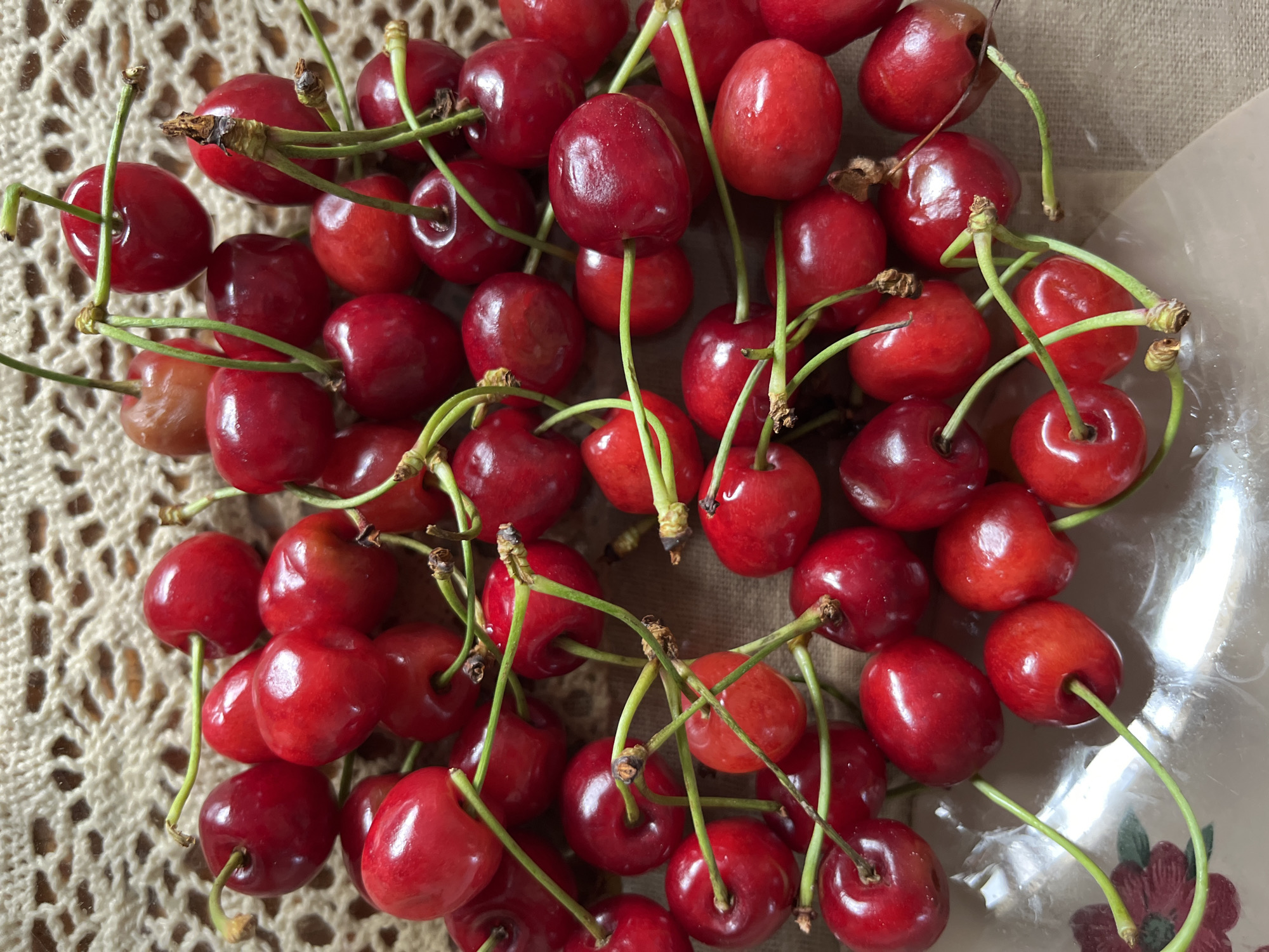 A pile of red cherries with green stems is scattered on a textured, beige surface, possibly a doily or woven fabric. Some cherries are clustered together, while others are more spread out. The lighting suggests an indoor setting, with highlights reflecting off the smooth surfaces of the fruit. No people or animals are present in the image. The location is unknown.