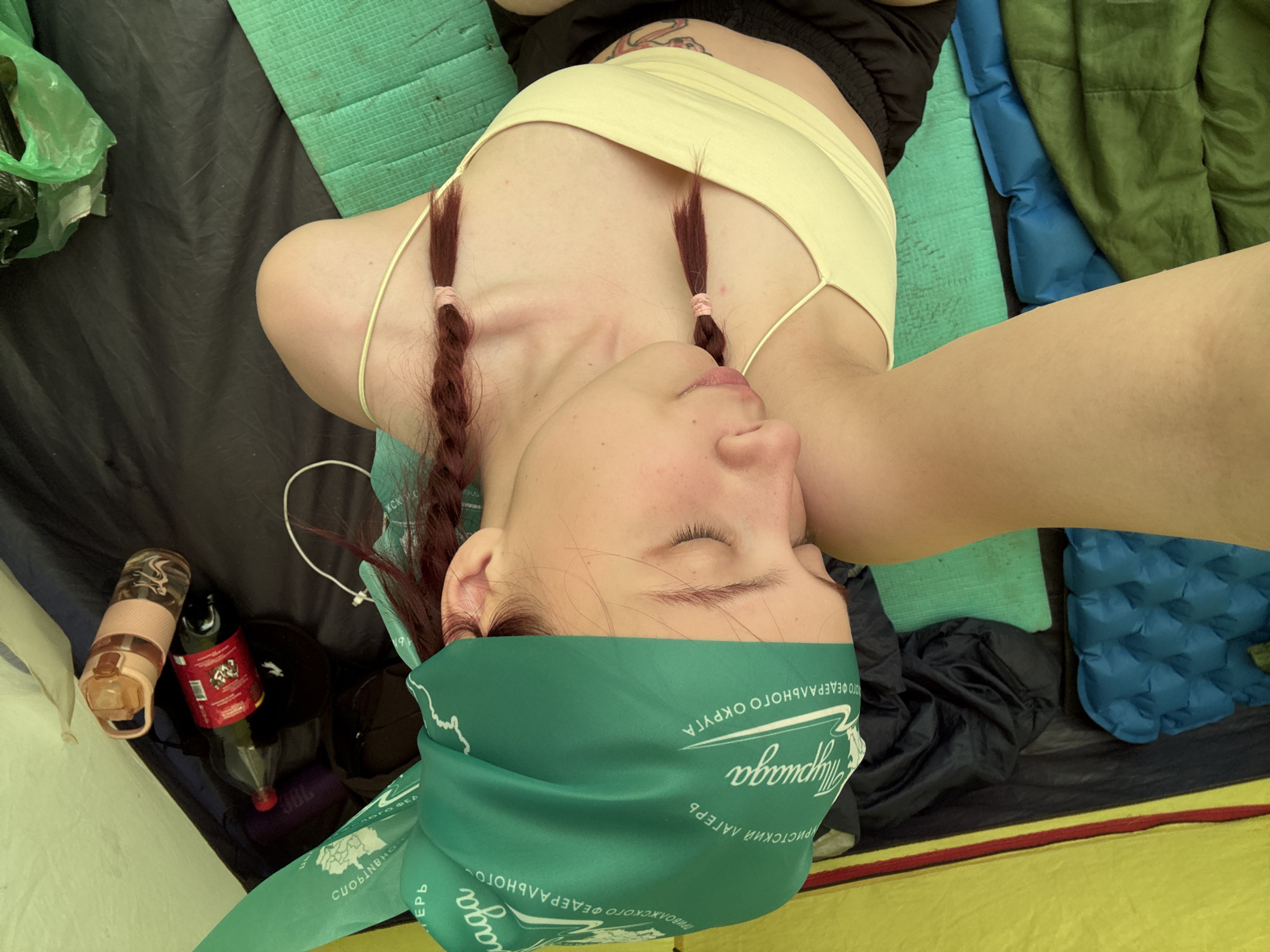 A person with auburn hair in small braids, wearing a green bandana with Cyrillic text, lies inside a tent. The person is wearing a light yellow top, and their midriff is visible. Near them are a water bottle, a soda bottle with a red label, and camping gear including a blue inflatable pad and a green sleeping bag. The floor of the tent is black with a yellow and red accent. The exact location in Khvalynsk, Russia cannot be determined from this image.