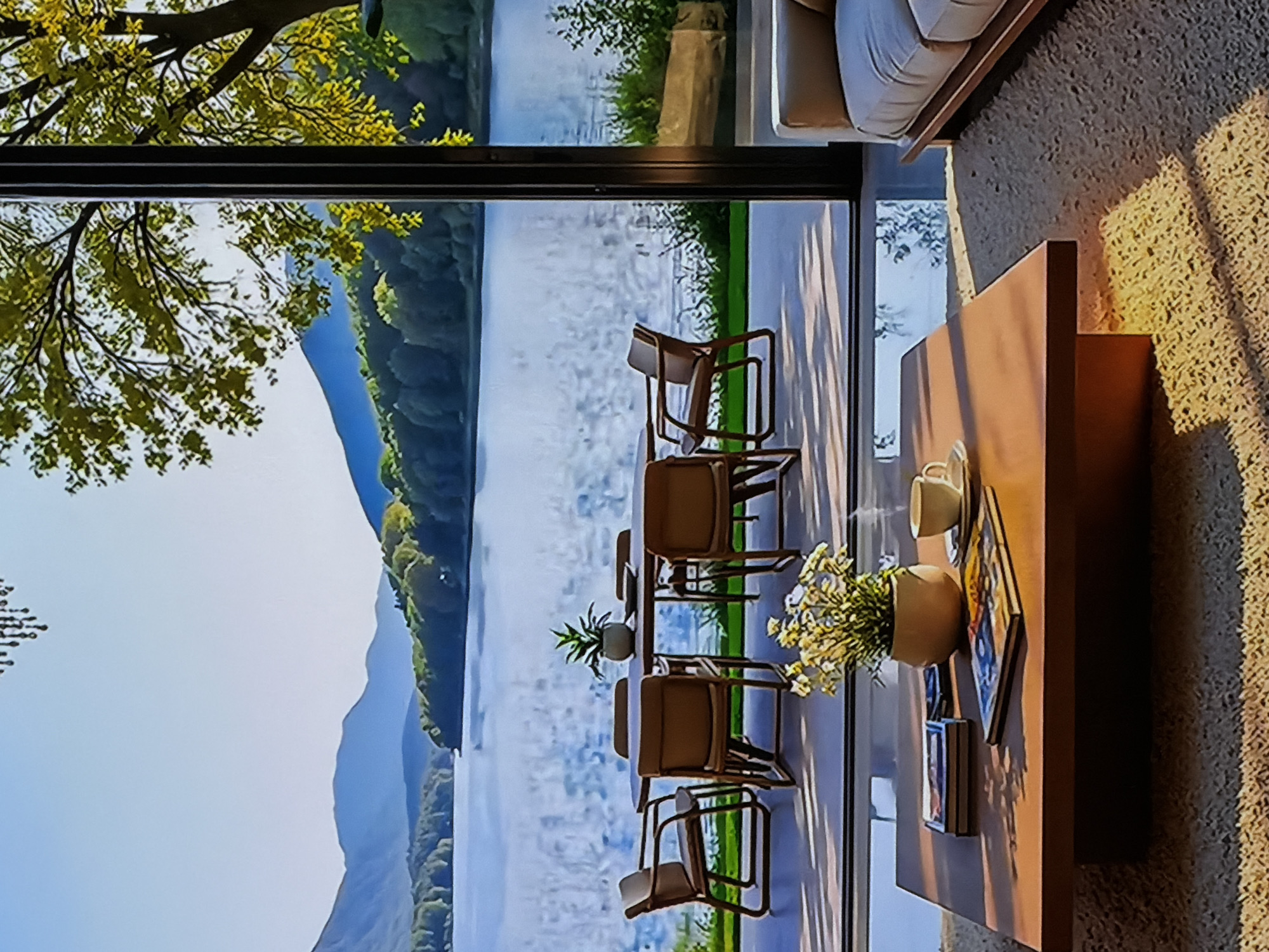 This media file captures a serene interior living space featuring a large glass window that offers a panoramic view of an outdoor natural landscape. The image is oriented vertically, showing the scene from a slightly elevated perspective.
In the indoor foreground on the right, a section of a light-colored sofa is visible, next to a textured, possibly carpeted, wall. A wooden coffee table is prominently placed in the lower right, adorned with a light-colored ceramic cup, a vase holding white and yellow flowers, and what appears to be books or magazines. The bright sun casts distinct shadows from these items onto the table and the adjacent wall, suggesting it is either morning or late afternoon.
Beyond the expansive window, an outdoor patio area is visible, furnished with a dining table and several brown chairs. A potted plant is centered on the outdoor table. The background unfolds into a breathtaking natural vista: a wide body of water, likely a lake or river, reflects the clear sky. Lush, green tree-covered mountains or hills line the distant shore, creating a tranquil backdrop. A large tree branch with vibrant green and yellow foliage extends into the upper left portion of the frame, framing the natural view.
No people are present in the scene, contributing to a sense of peacefulness and calm. The clear lighting indicates sunny weather. The setting suggests a modern dwelling designed to maximize integration with its natural surroundings. There is no visible text in the image. While the provided location context is Sofiivska Borshchahivka, Ukraine, the image itself does not contain specific visual cues to confirm this geographical location.